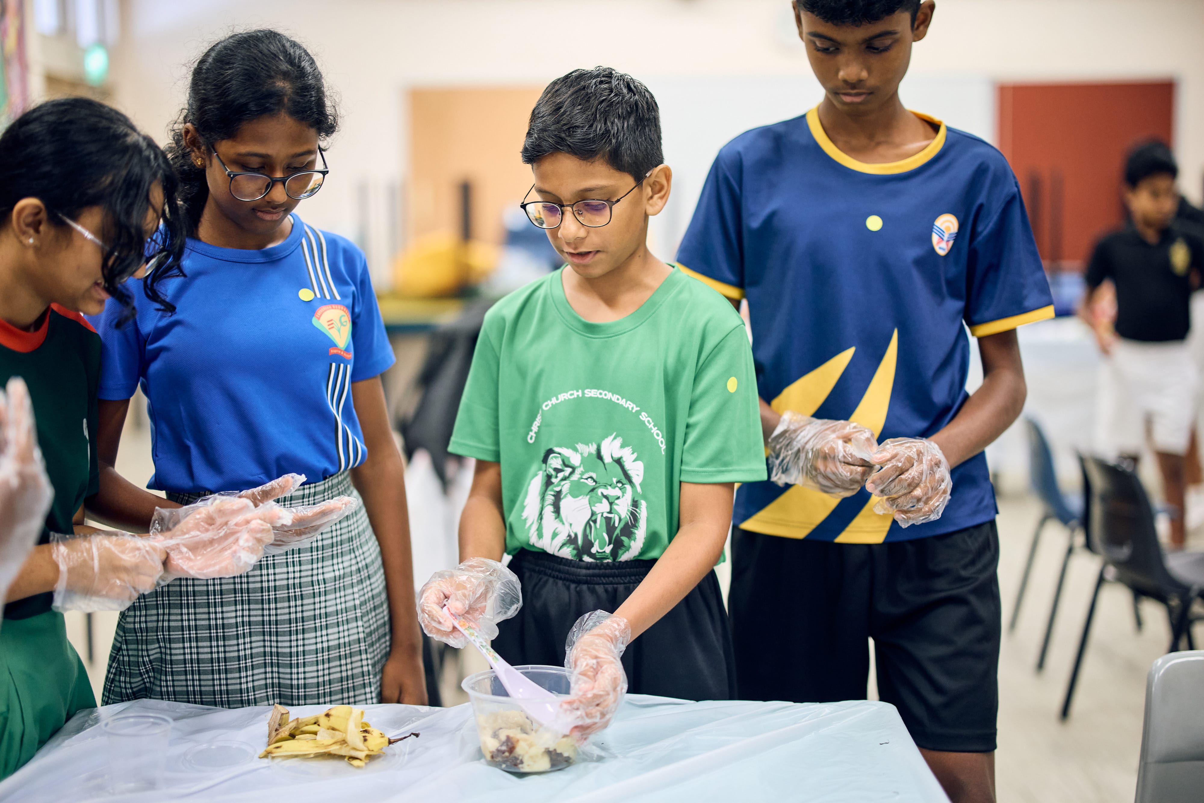 Four students wearing gloves stand at a table, mixing food scraps in a clear plastic container.
