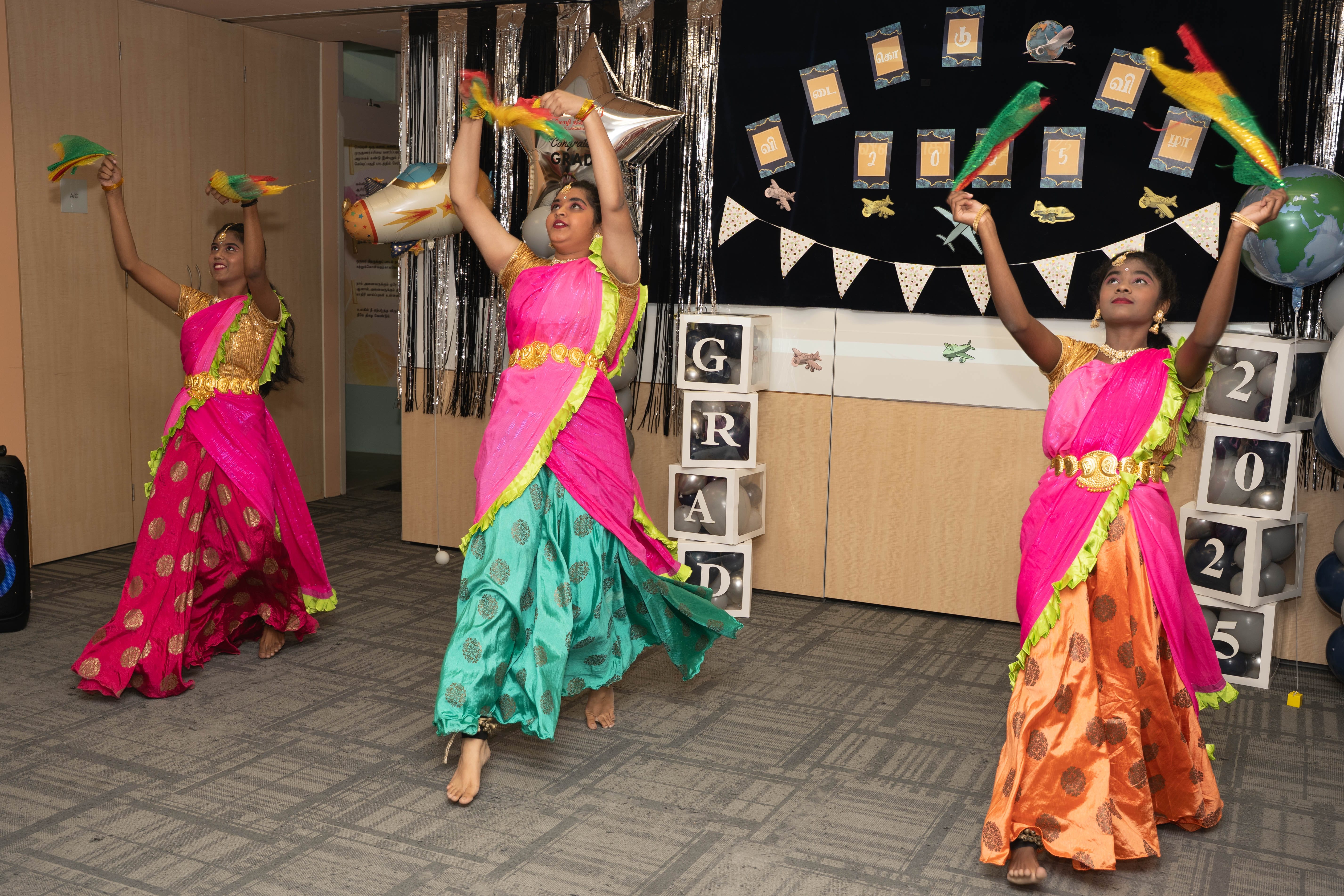Three dancers in bright pink saris raise feathered fans on a decorated indoor stage with balloons.