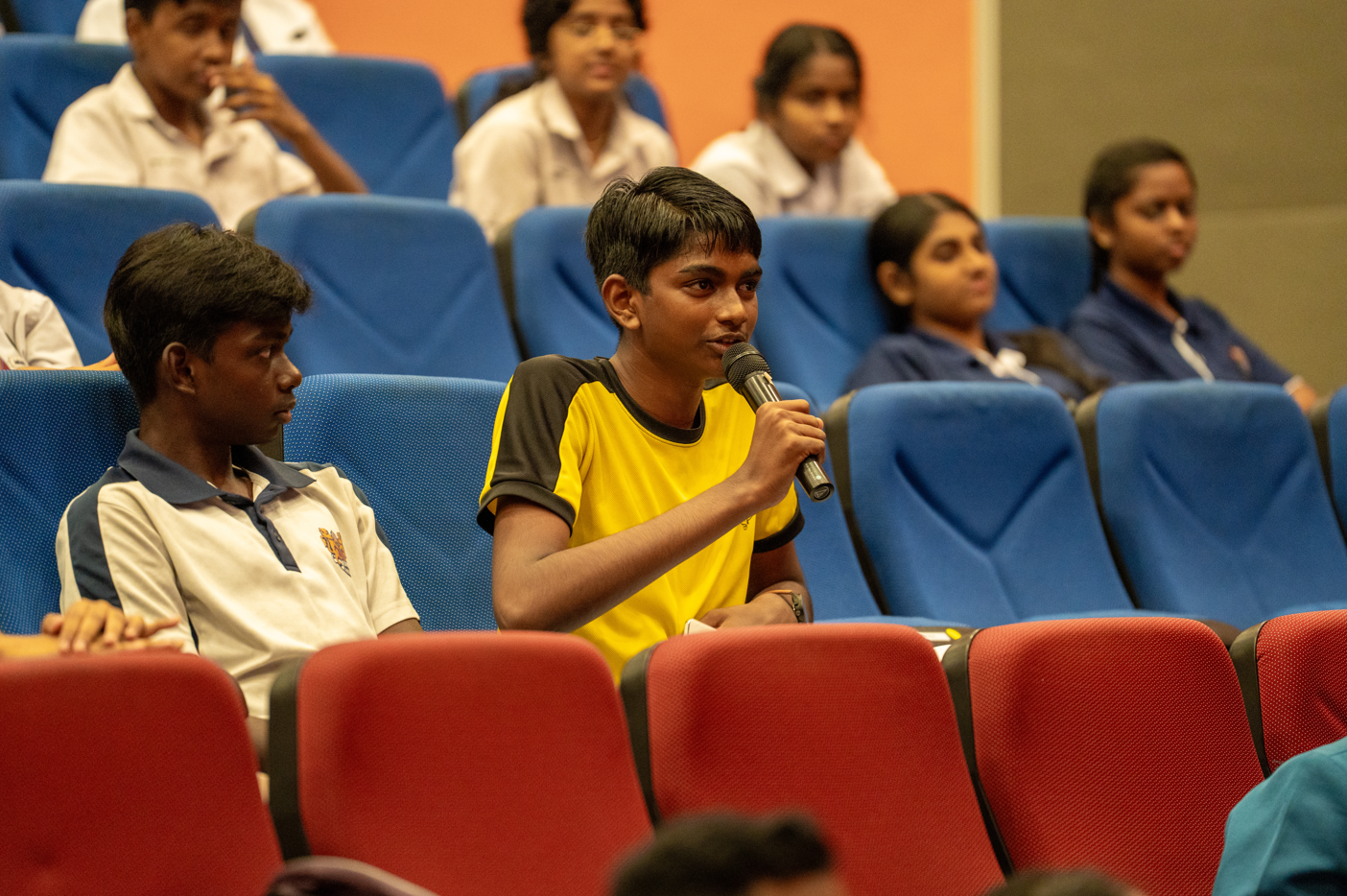 Student in yellow shirt speaks into a microphone while seated among classmates in blue auditorium seats
