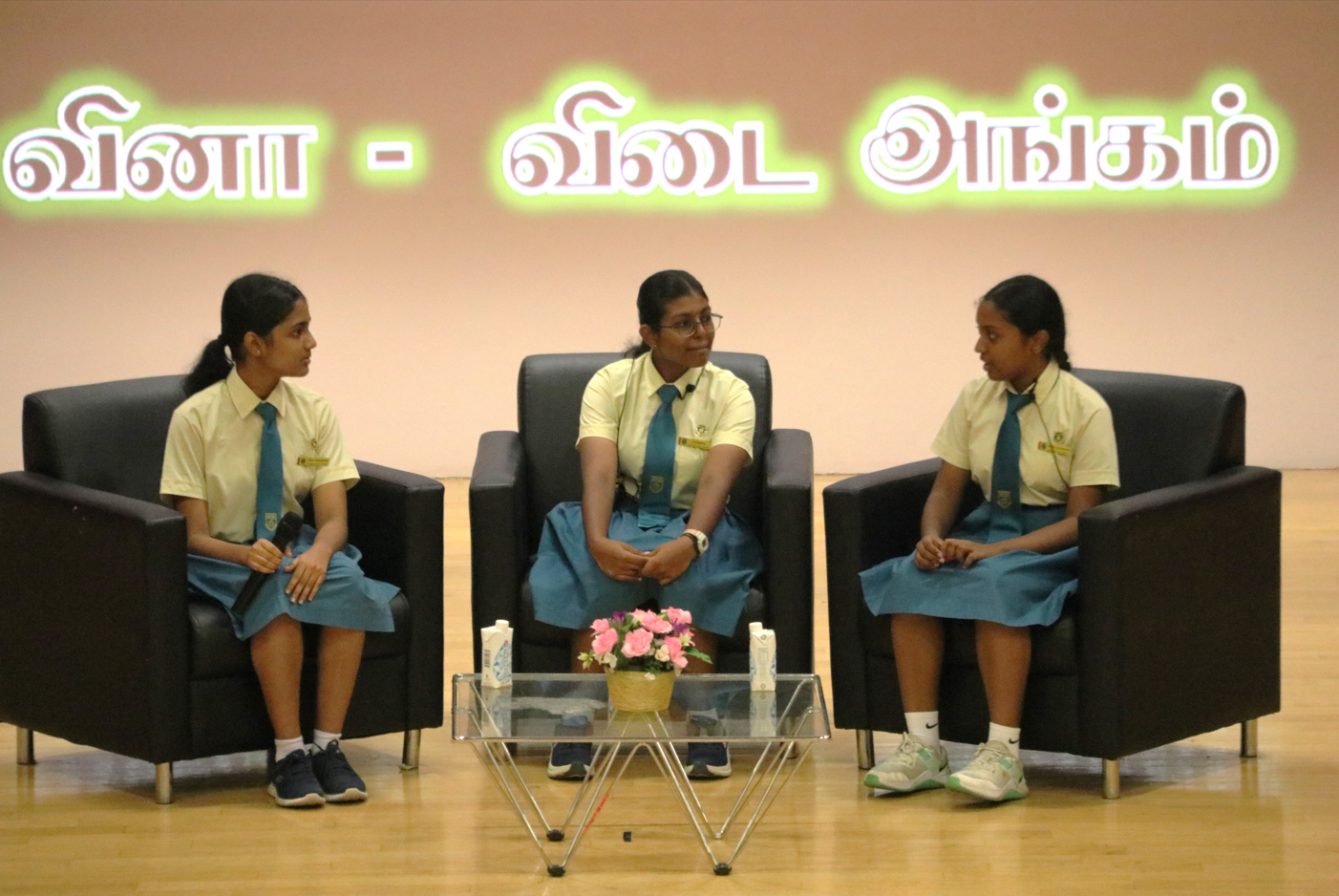 Three schoolgirls in uniform sit in black armchairs onstage, with Tamil text glowing behind them.