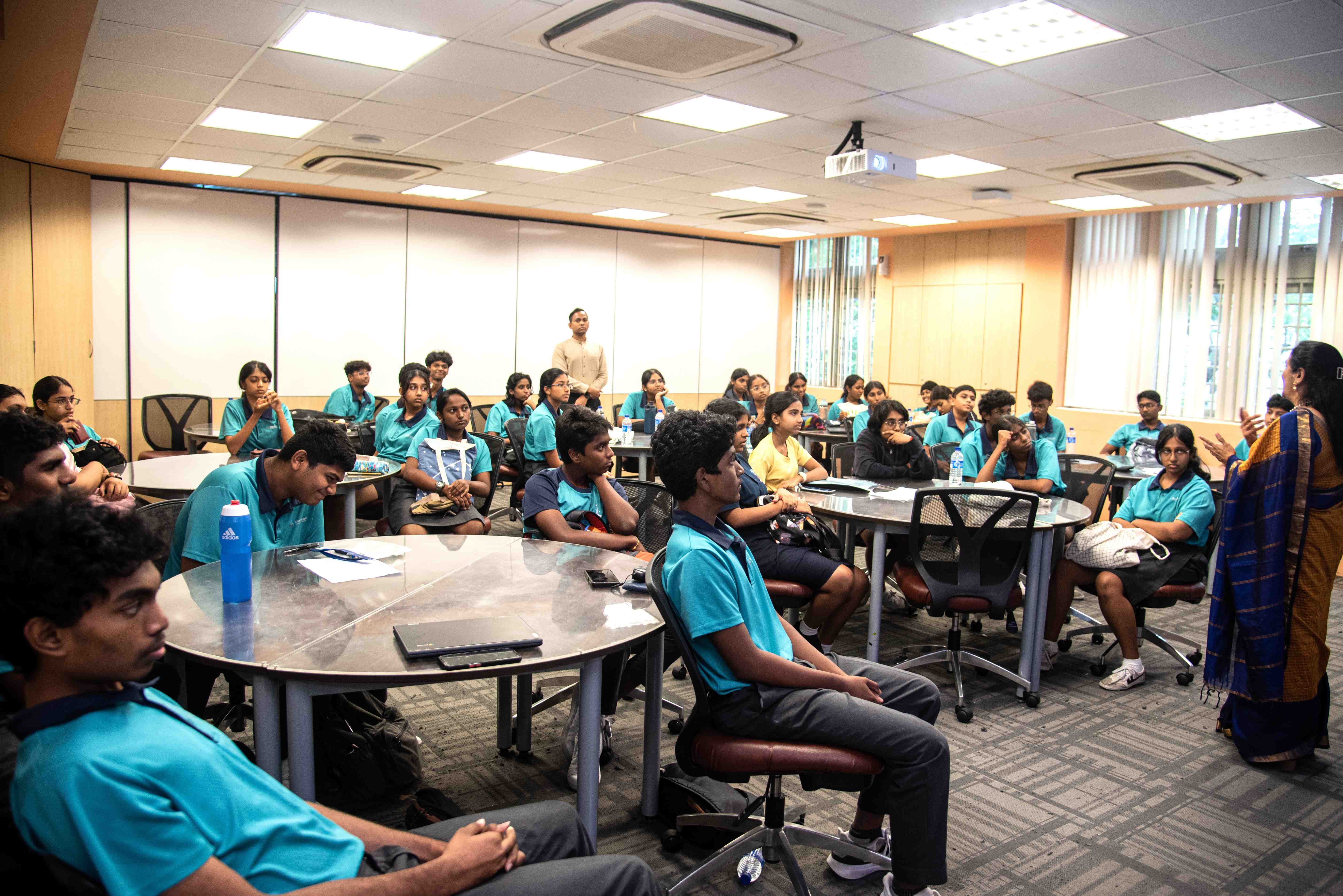 Students in teal uniforms sit at round tables listening to a woman speaking in a classroom.