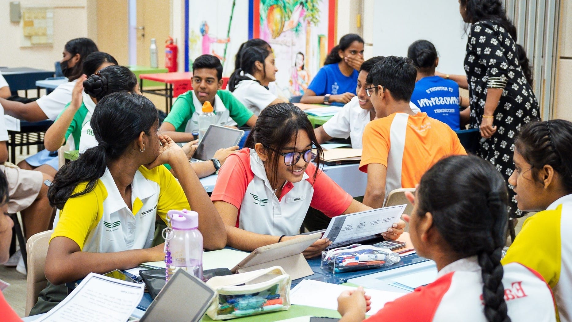 Students in uniforms sit at tables, talking and reading papers, as a teacher stands nearby.