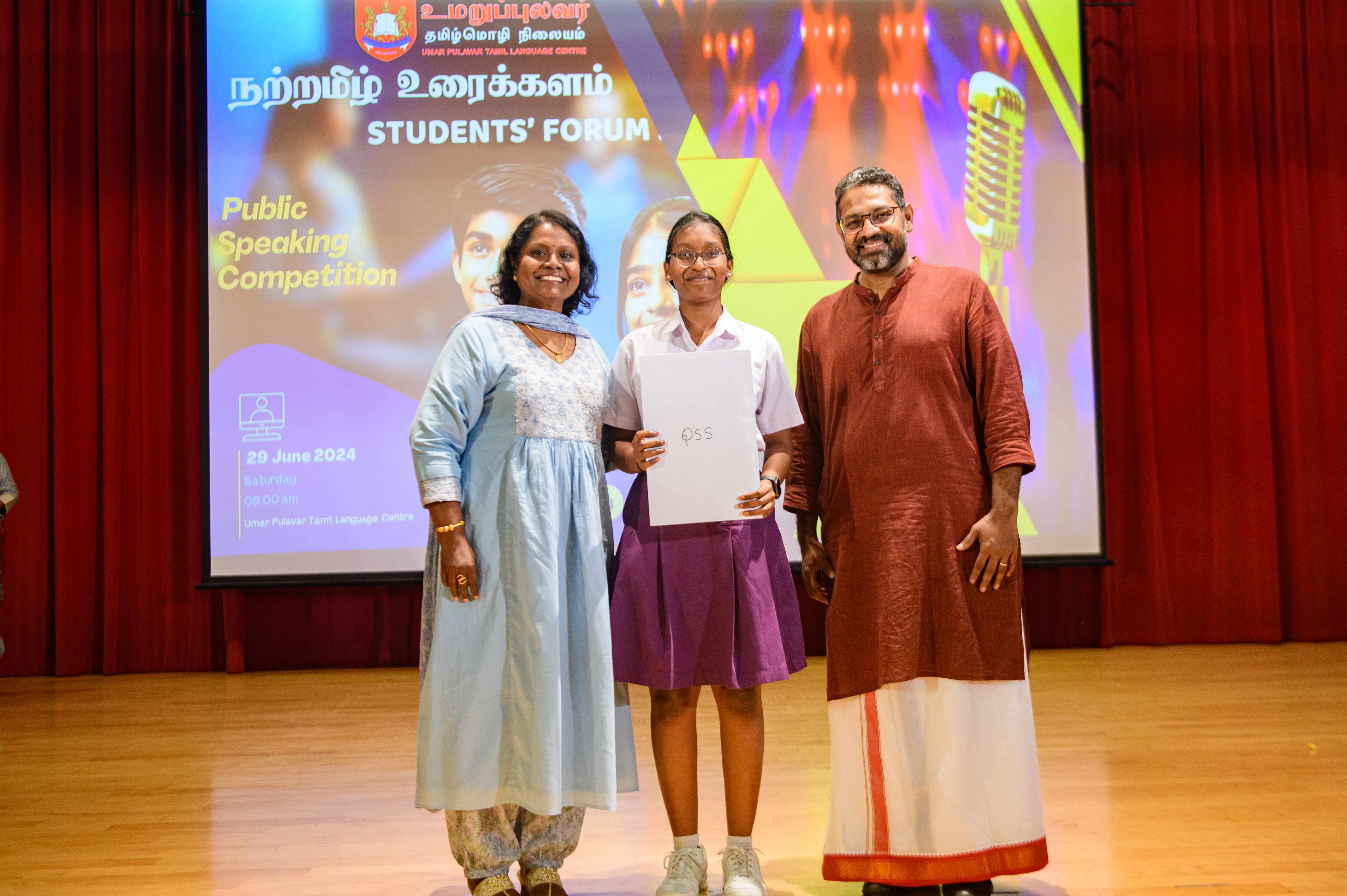 Three people on stage; student holds a certificate in front of a Students’ Forum public speaking backdrop.