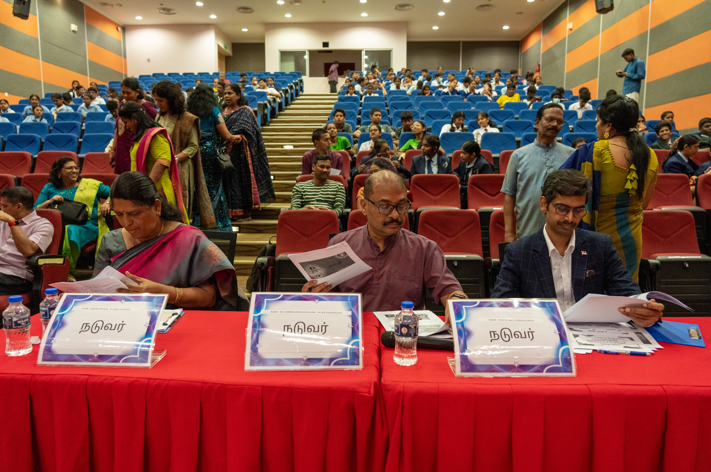 Three judges at a red table read papers in an auditorium with many seated attendees behind them.