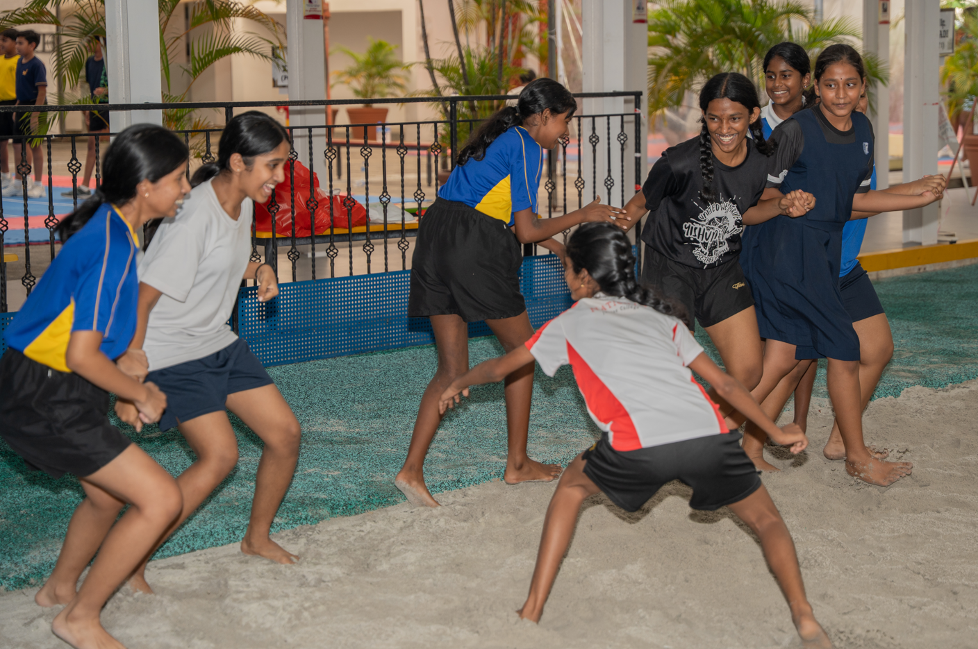 Group of girls laughing and playing a team game barefoot on sand in a covered courtyard.