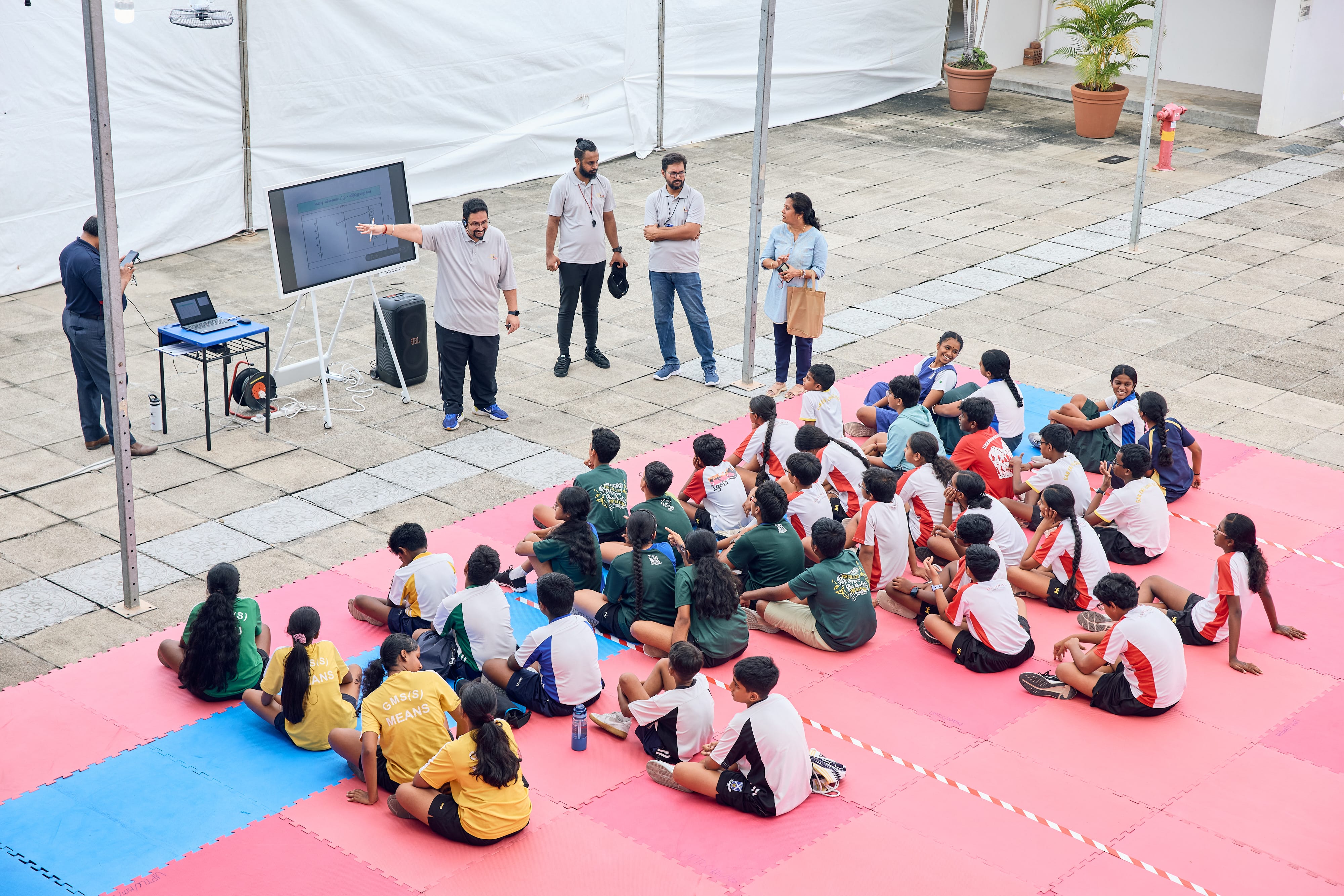 Man speaks by a screen while students sit on red and blue mats in an outdoor courtyard.