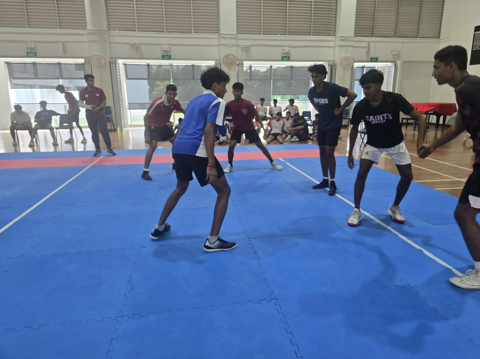 Players in sportswear face off on a blue indoor court while others watch from the back.
