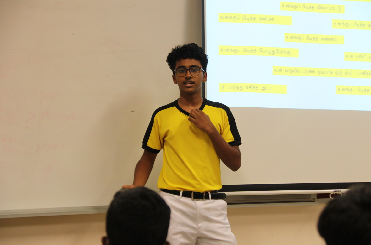 Student in yellow shirt and glasses presents in class beside a screen with highlighted Tamil text.