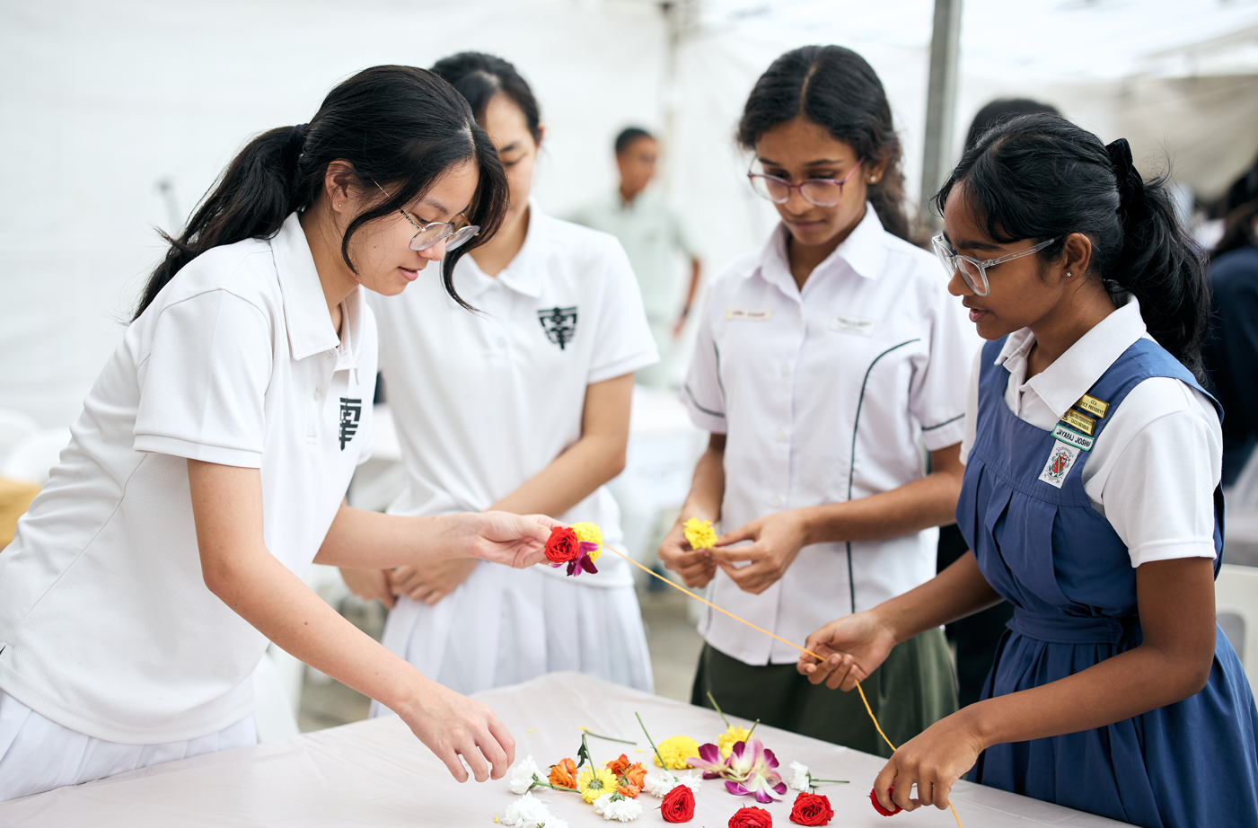 Four students in school uniforms arrange colorful flowers on a table, threading a garland together.