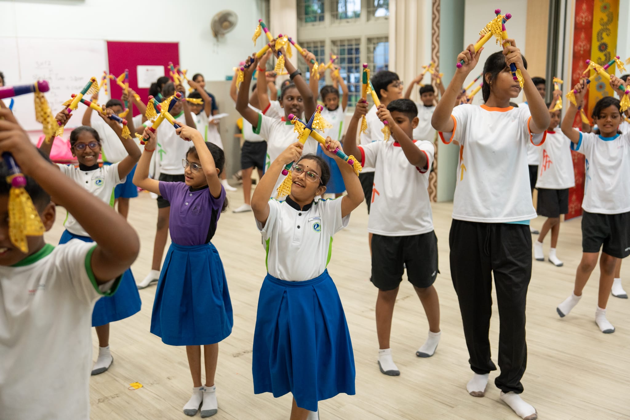 Students in a hall raise colorful sticks overhead during a coordinated activity, some in uniforms and blue skirts.