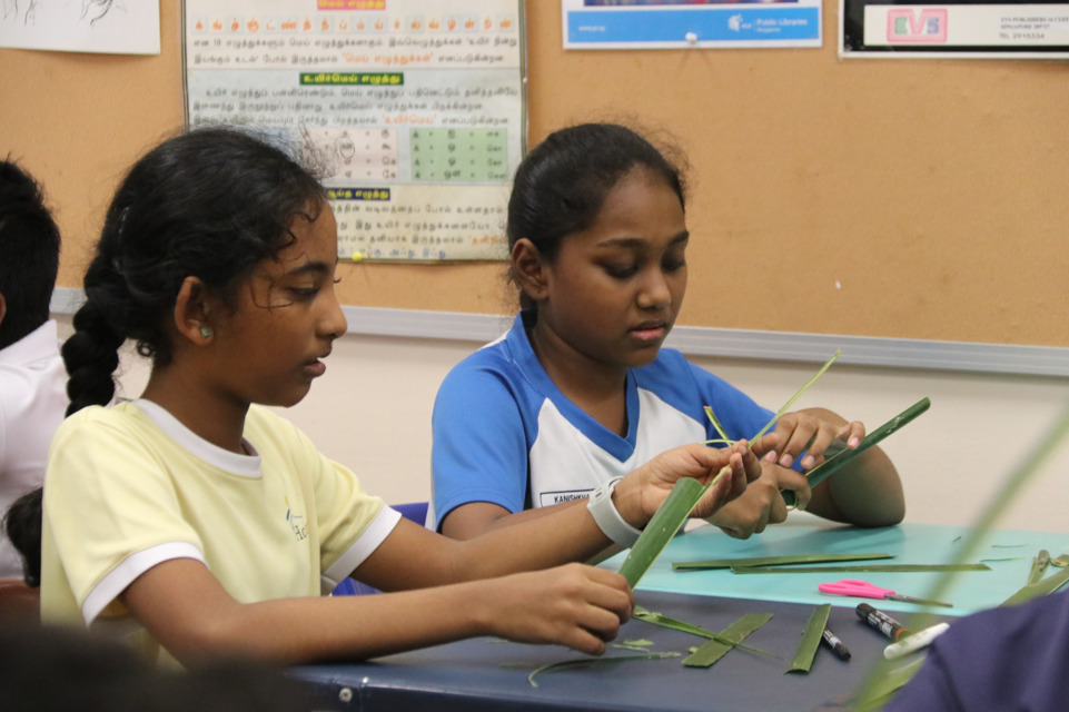 Two girls sit at a classroom table, handling green leaves with scissors and markers nearby.