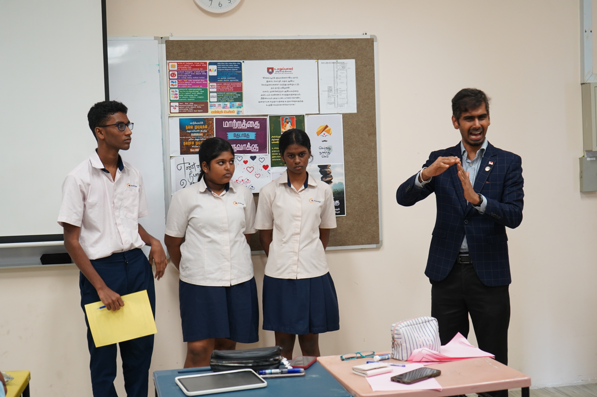 Teacher in blazer gestures while three uniformed students stand by a bulletin board with posters.