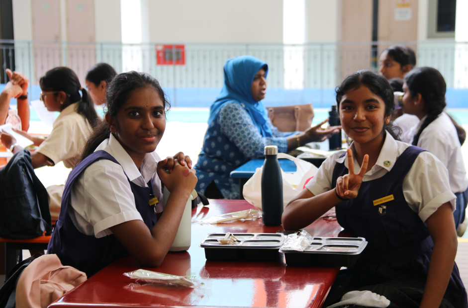 Two schoolgirls in uniforms sit at a cafeteria table; one smiles and flashes a peace sign.