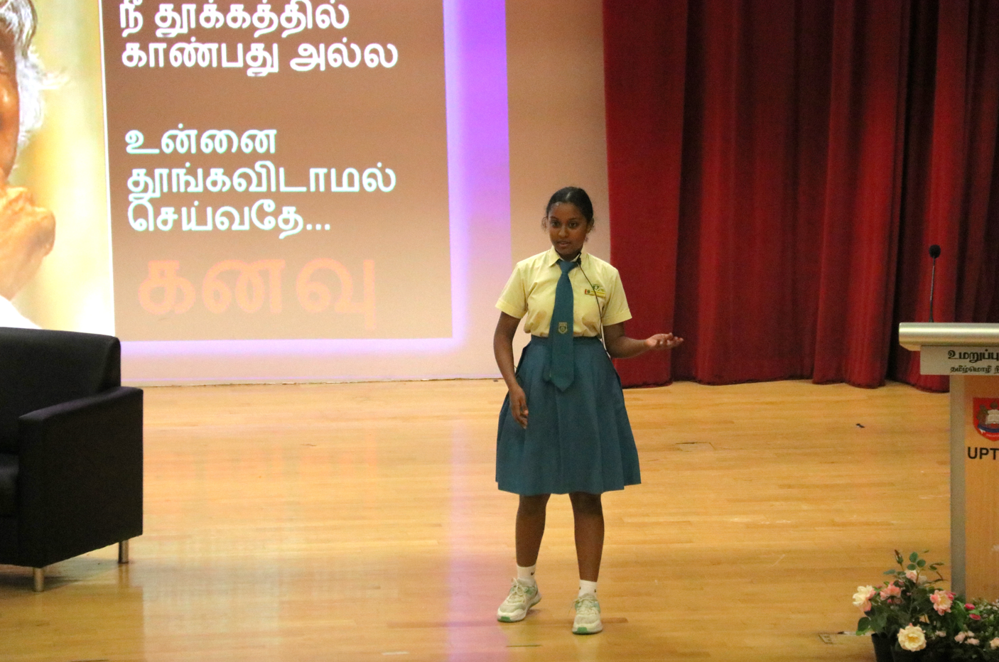 Schoolgirl in uniform speaks on stage beside a podium, with Tamil text projected behind and red curtains.