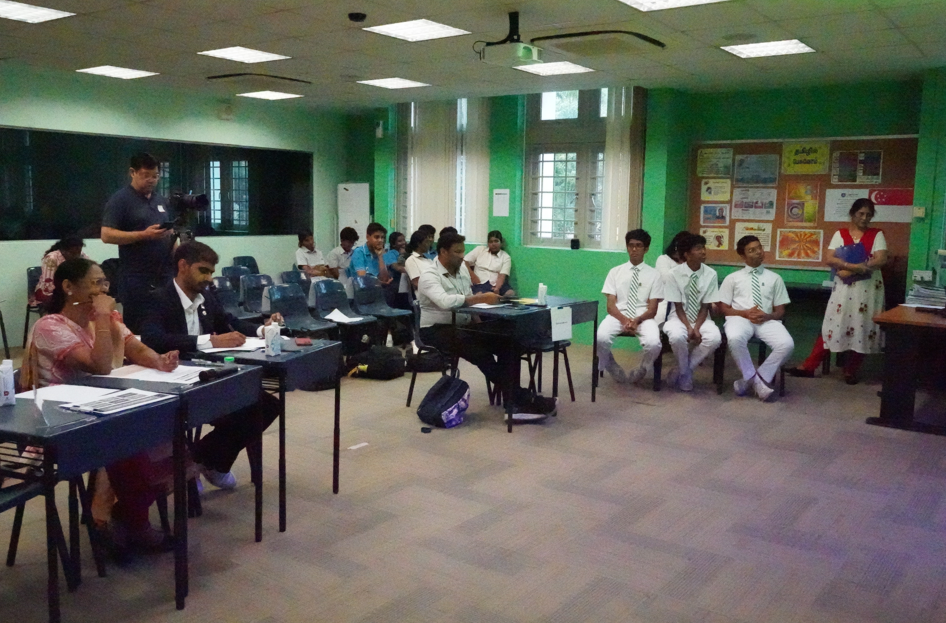 Classroom scene with seated students, judges at desks, and a woman standing by a bulletin board.