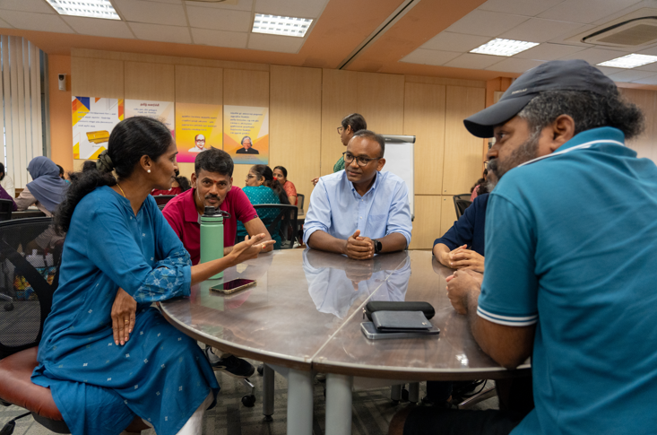 Four people sit around a round table in an office, talking; a water bottle and phones on the table.