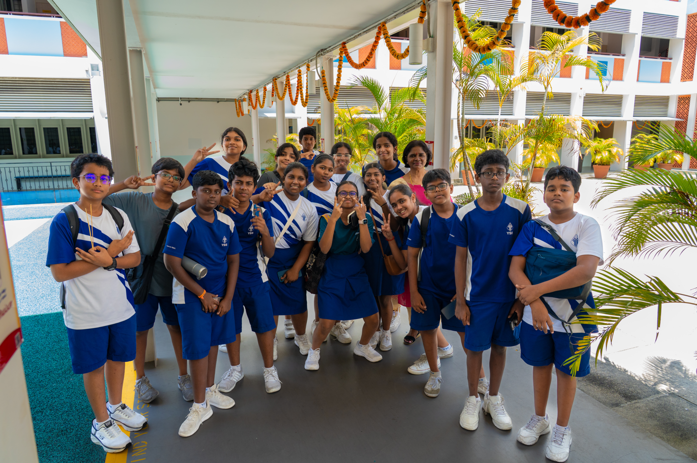 Group of students in blue uniforms pose under a decorated walkway, with palms and school buildings behind.
