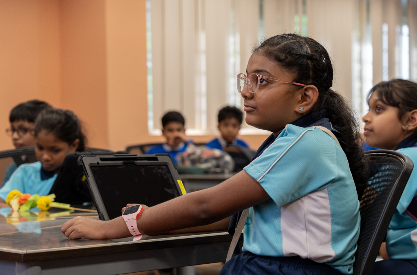 Girl with glasses sits at a classroom desk beside a tablet, while other students sit behind her.