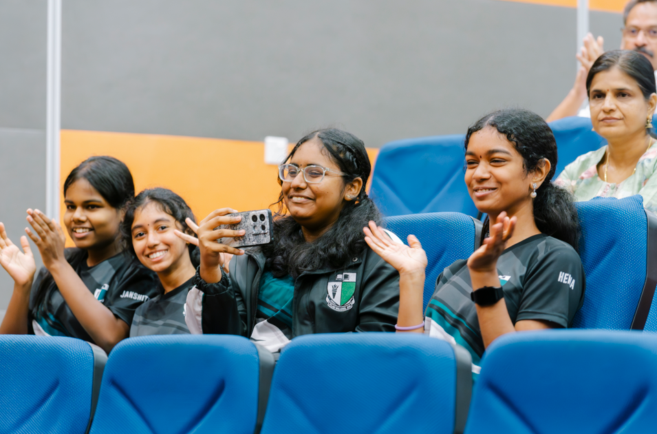 Four young women seated in blue chairs clap and smile; one holds up a phone to record.