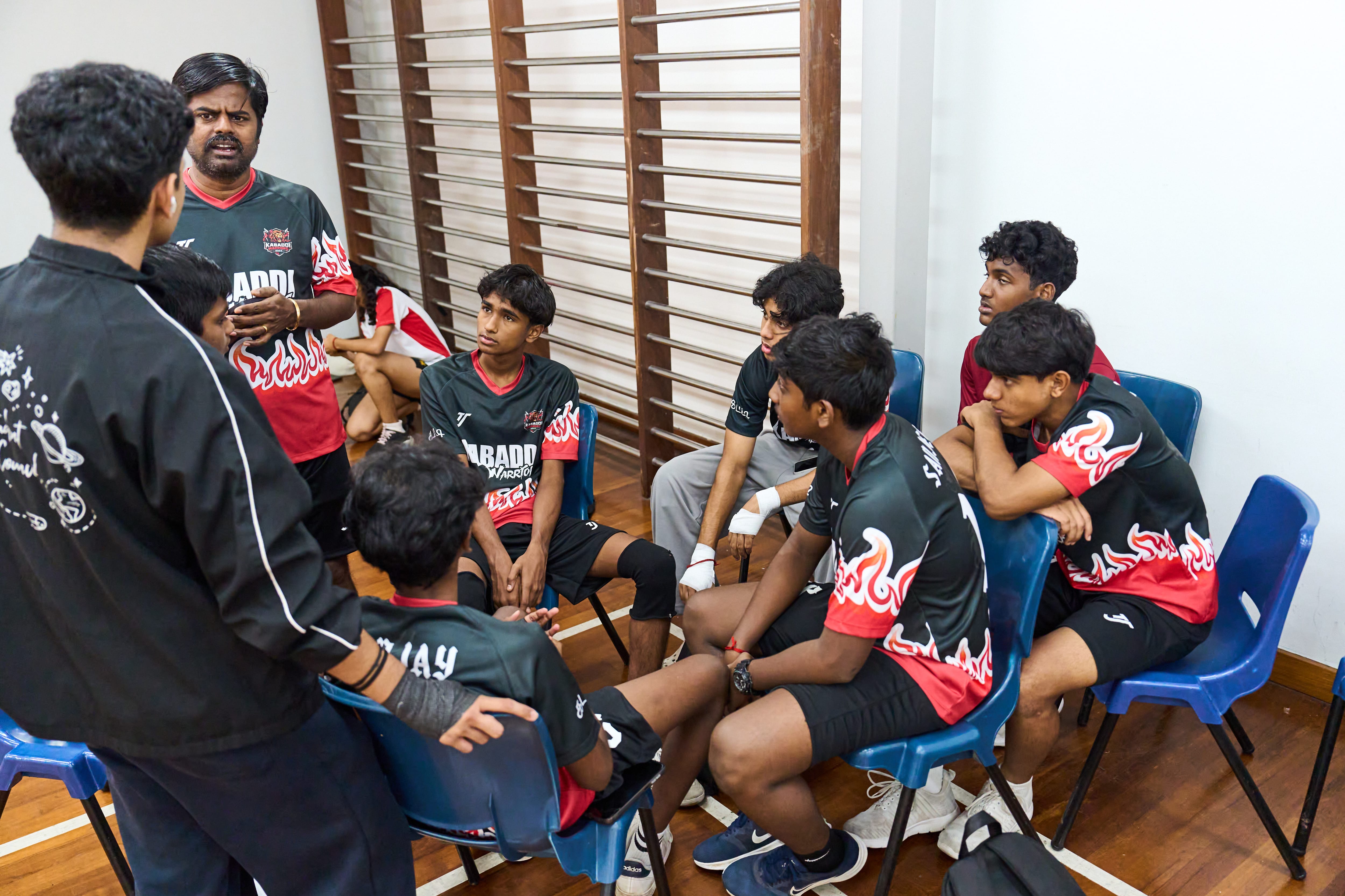 Coach speaks to seated players in black-and-red uniforms in a room with wooden wall bars and blue chairs.