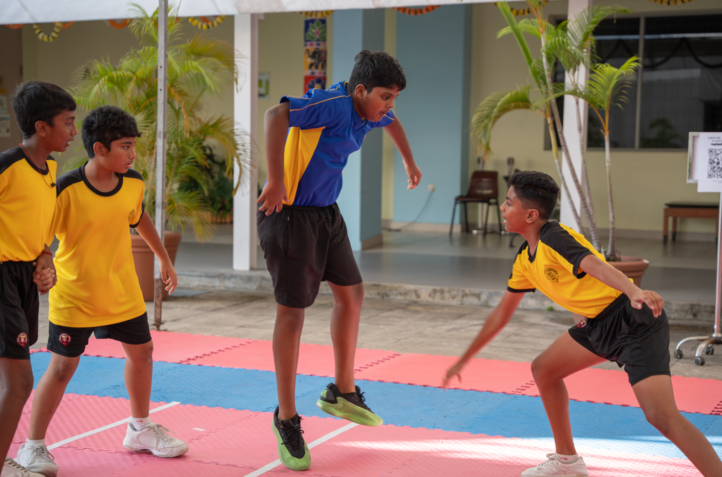 Boy in blue jumps on mats as three boys in yellow uniforms face him in a school courtyard.