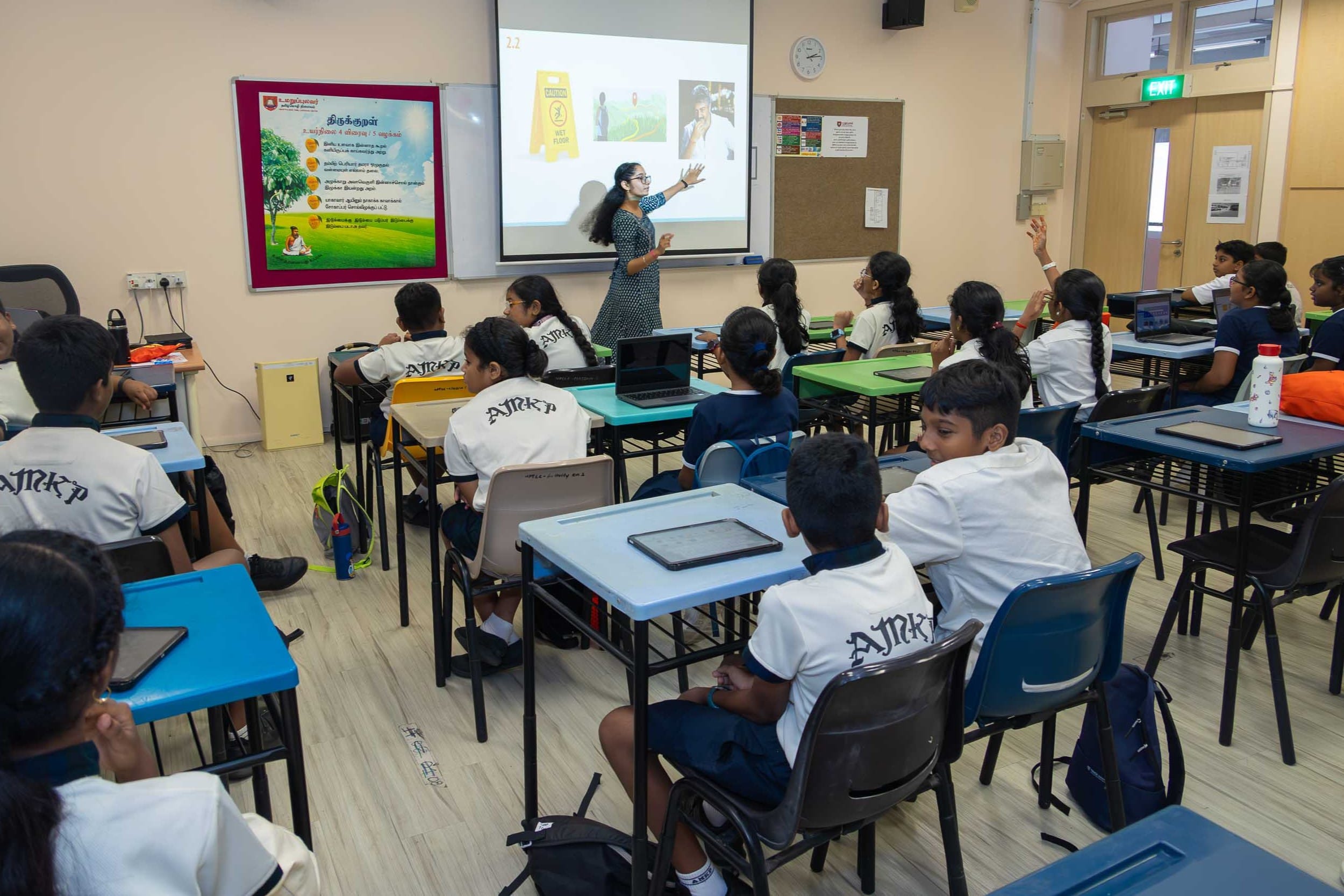 Teacher points to a projected slide while students in uniform sit at desks, some raising hands.