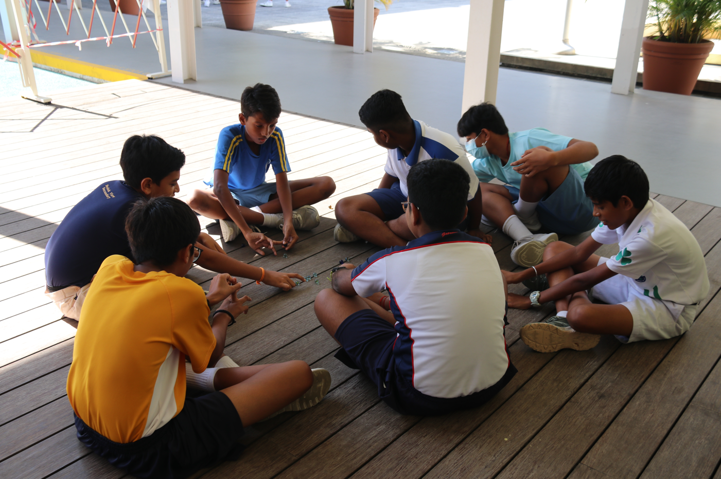 Seven boys sit in a circle on a wooden deck, playing a small game; one boy wears a face mask.