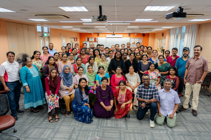 Large group of around 60 adults posing together in a room with an orange wall and ceiling projector.