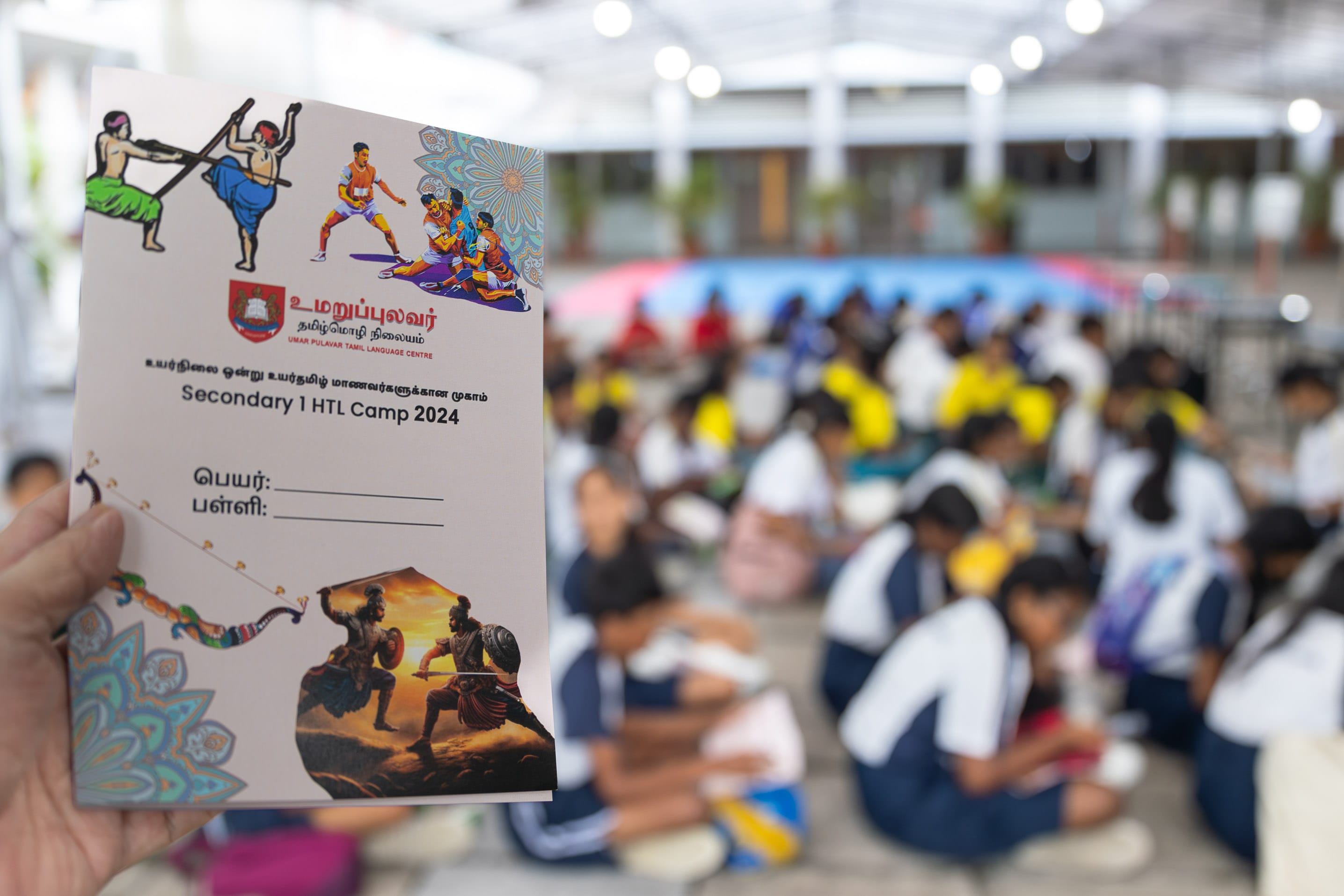 Hand holds a camp booklet; behind, blurred students sit on the floor in a bright hall.