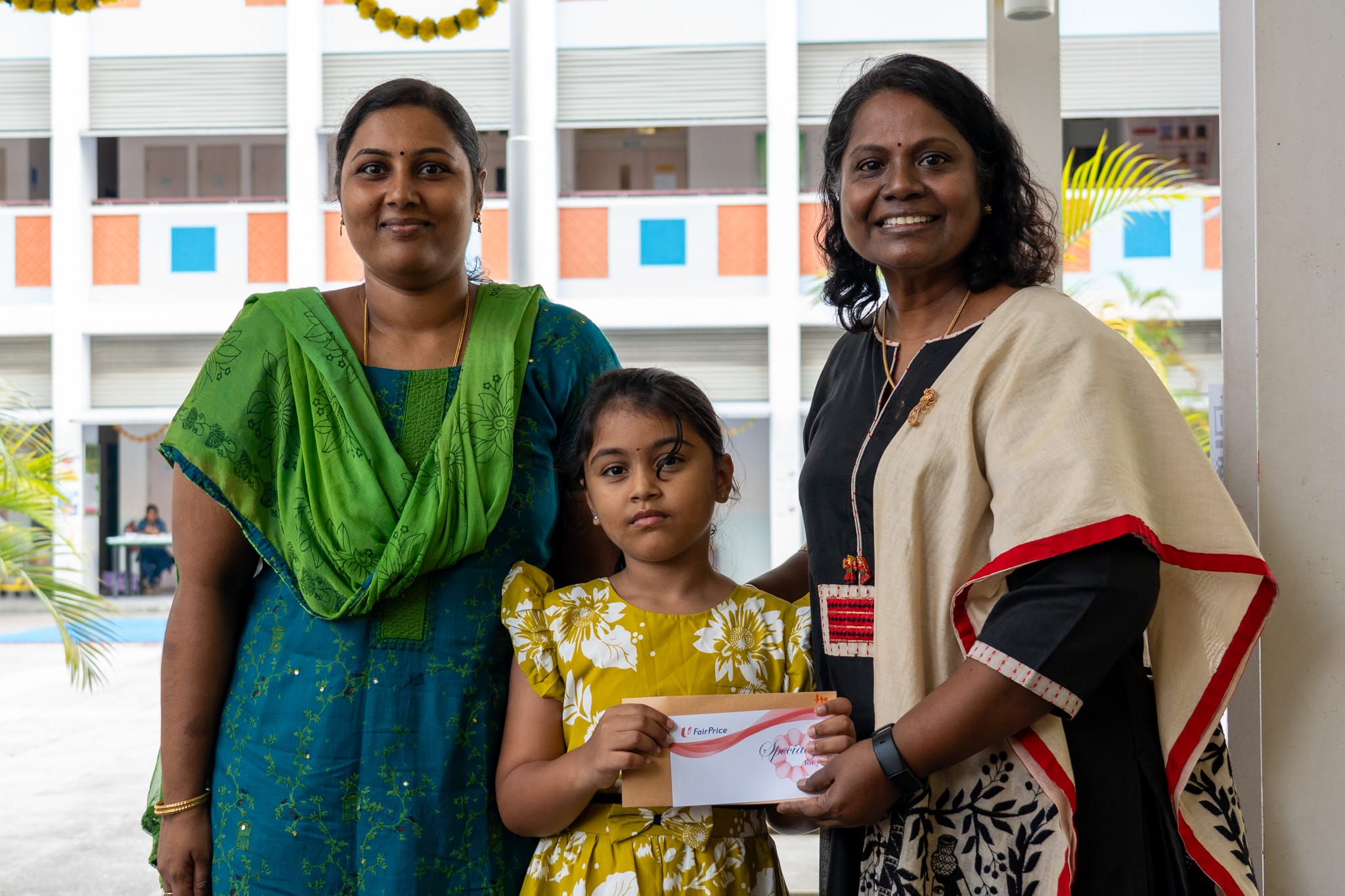 Two women stand with a girl holding an envelope, in front of a school building corridor.