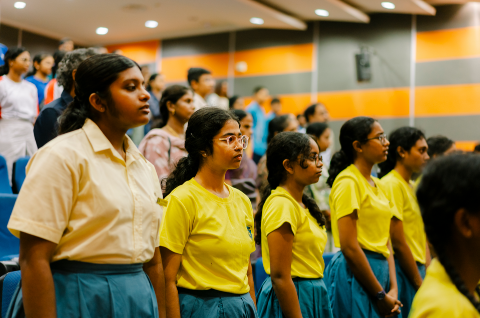 Students in yellow shirts and blue skirts stand in rows in an auditorium with orange and gray walls.