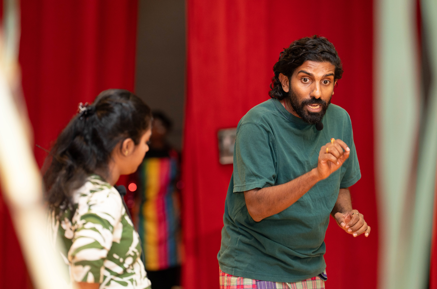 Bearded man in green shirt gestures while speaking to a woman in front of red curtains.
