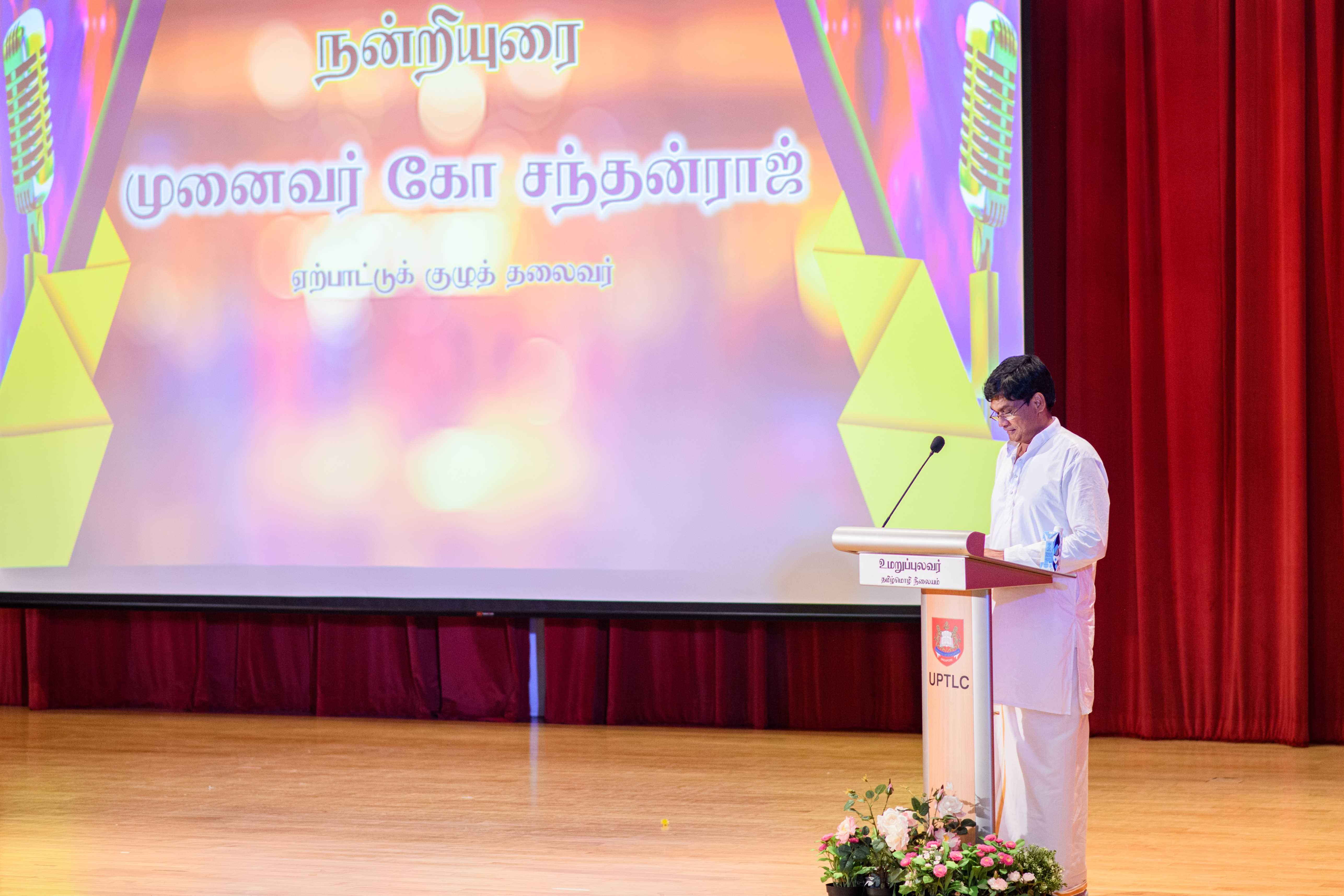 Man in white stands at a podium on stage, speaking before a large screen with Tamil text and microphones.