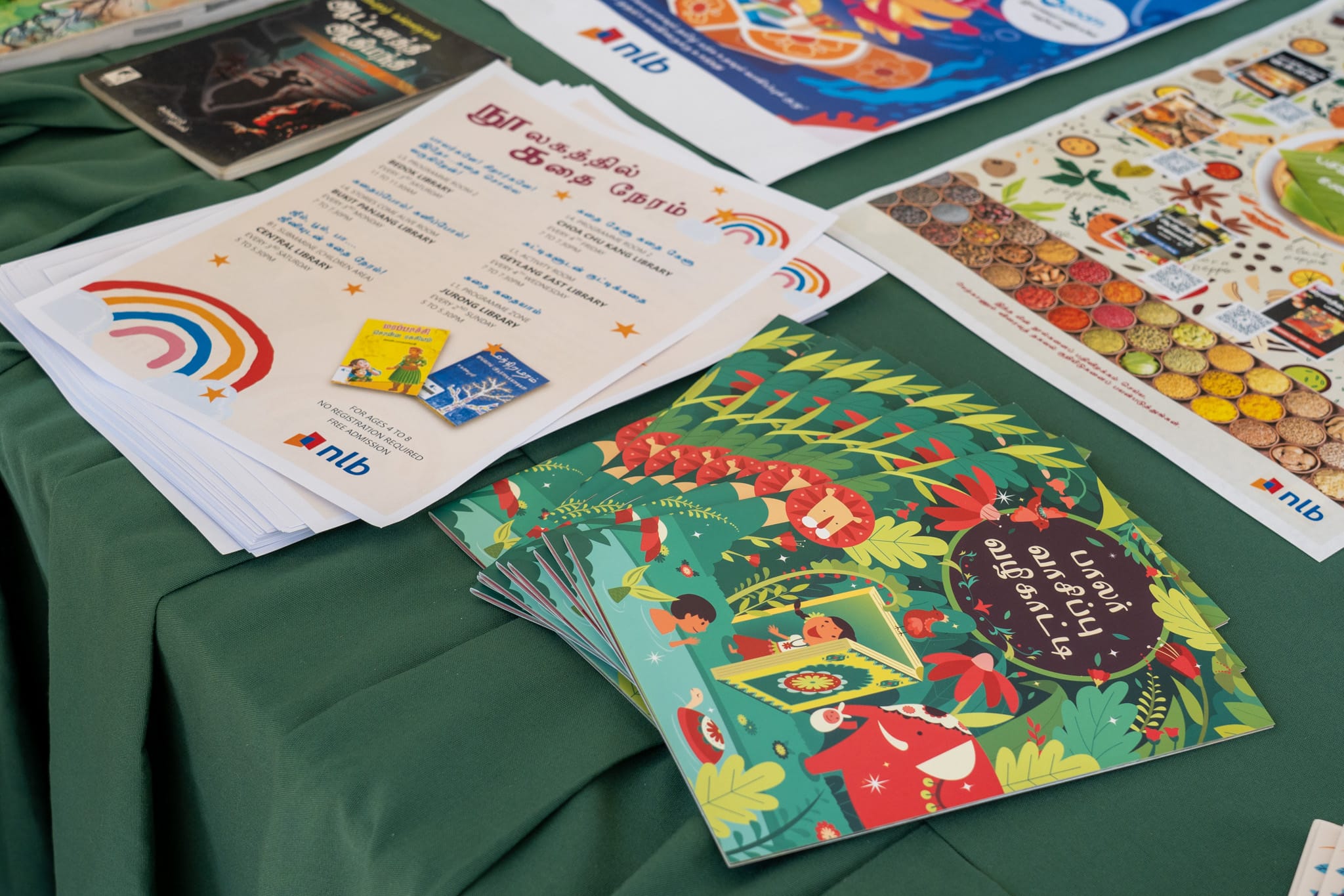 Stacked leaflets and colorful booklets with Tamil text and rainbow logos on a green tablecloth
