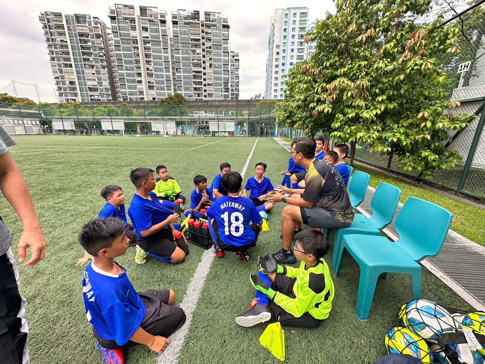 A coach talks to young soccer players in blue Waterway jerseys seated on a green turf field.