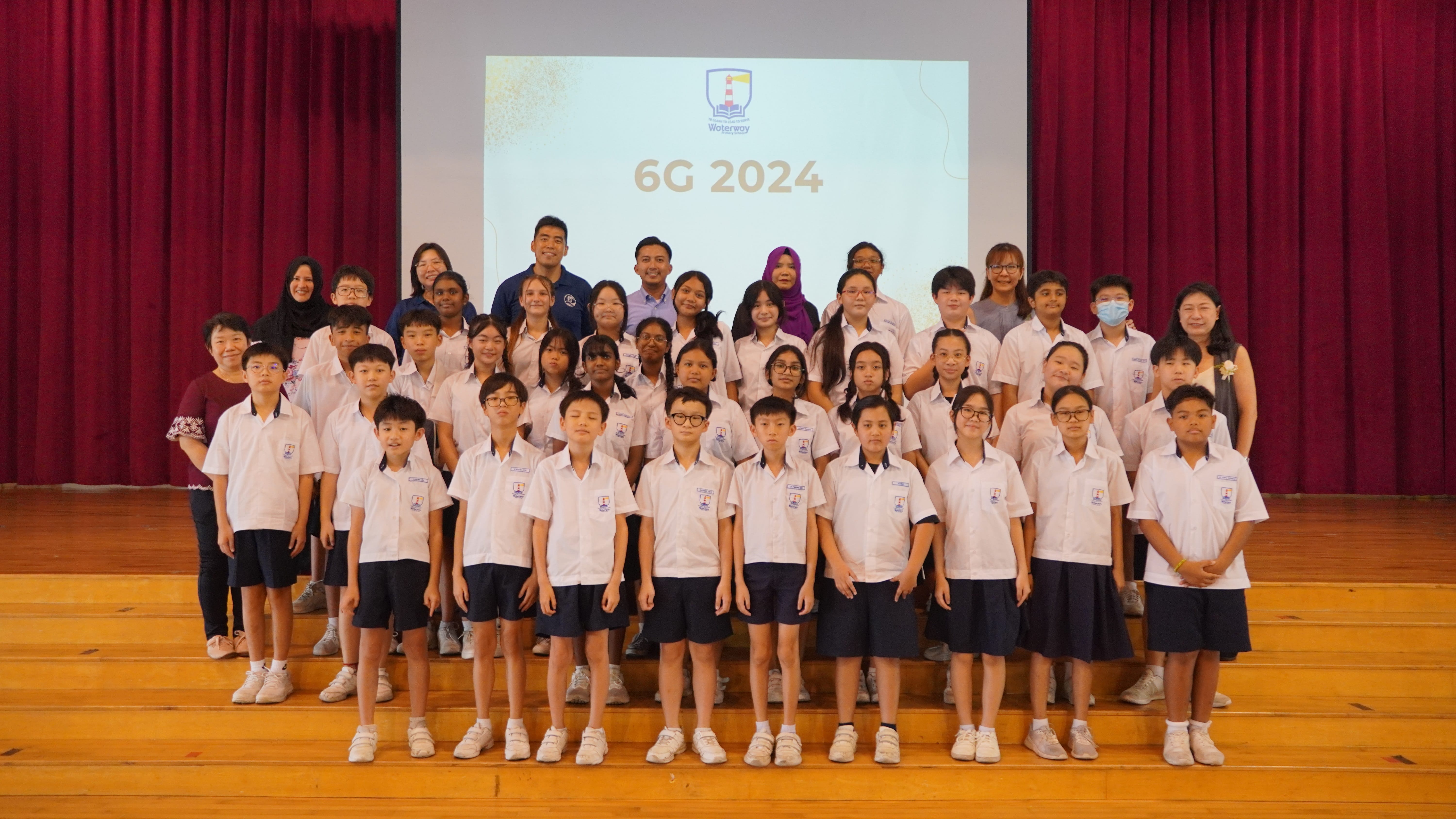 Group of students and teachers in white uniforms on stage; screen behind reads 6G 2024 with red curtains.
