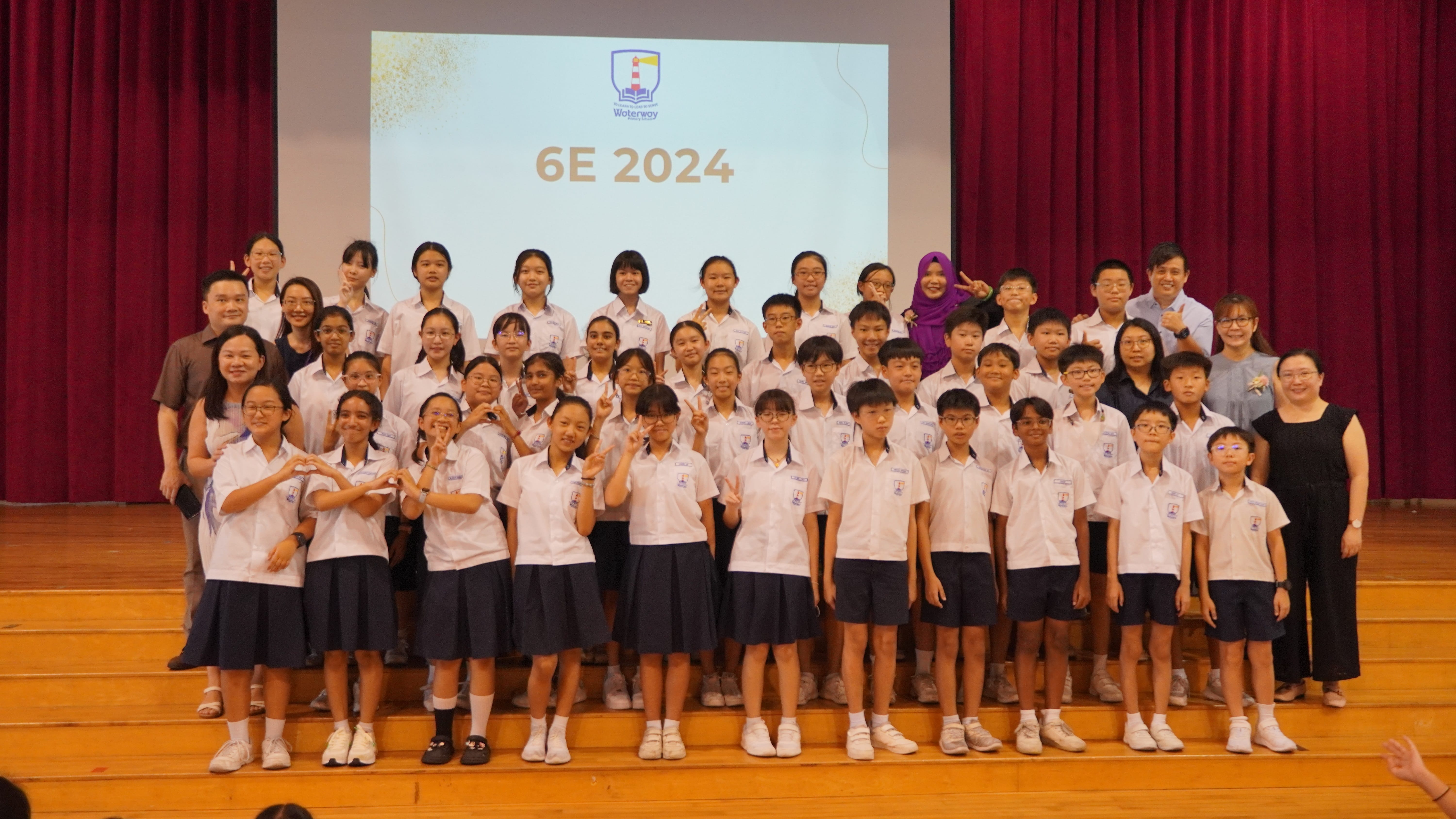 Large group of students and teachers posing on stage; screen behind reads 6E 2024.