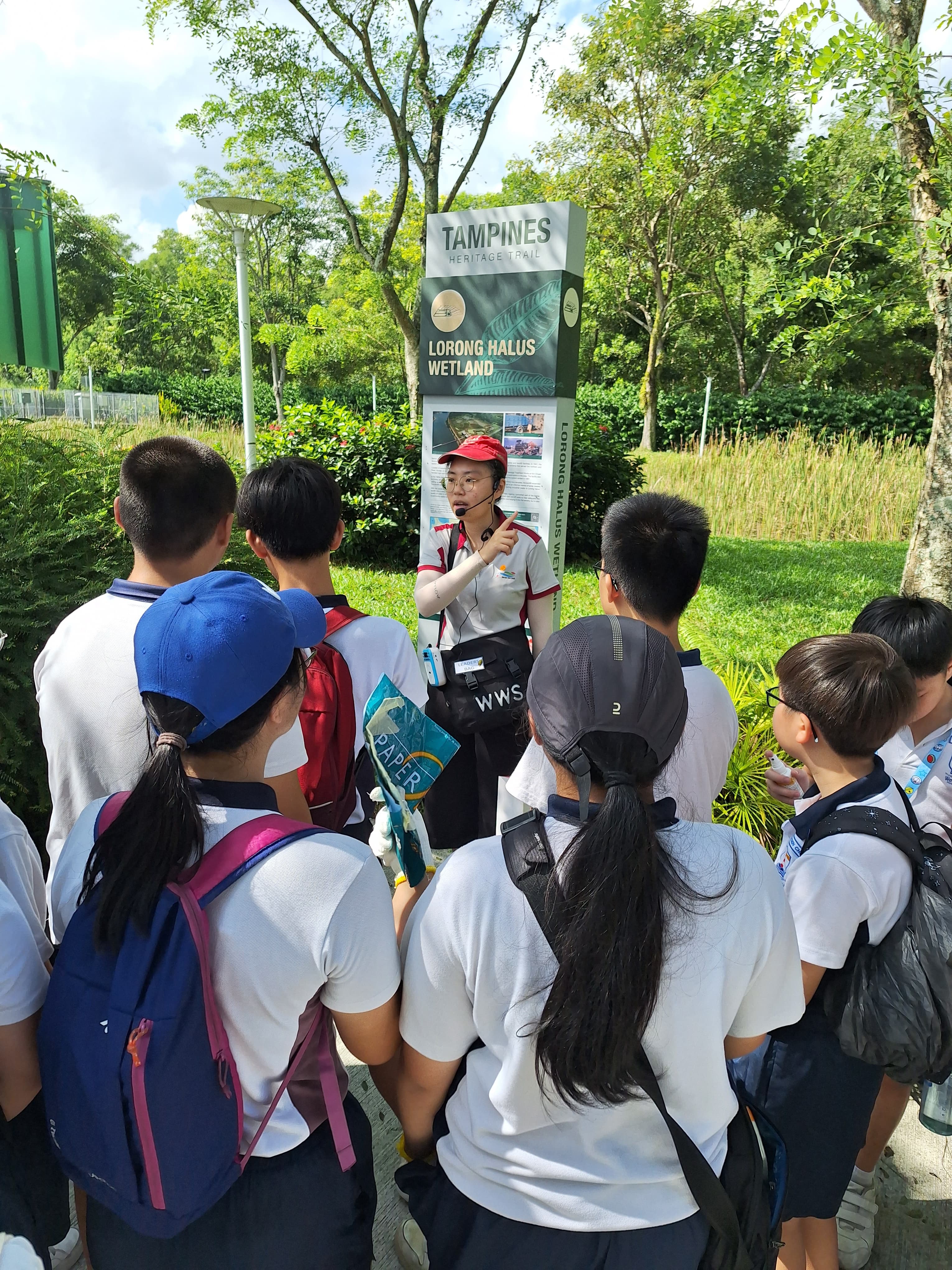 A guide speaks to a group of students at the Tampines Heritage Trail Lorong Halus Wetland sign outdoors.