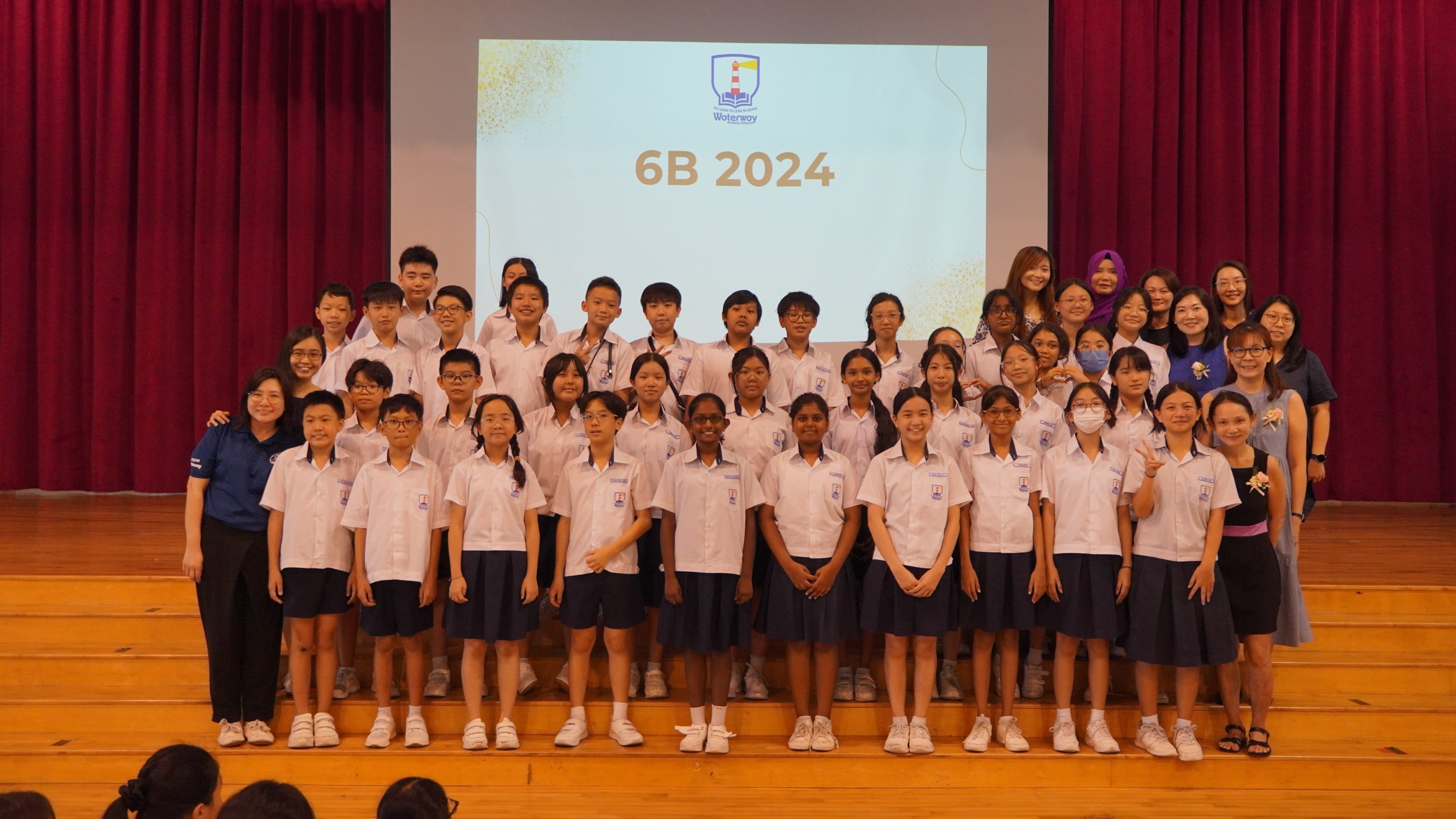 Group of students and teachers in uniform on a stage; projection behind reads 6B 2024.