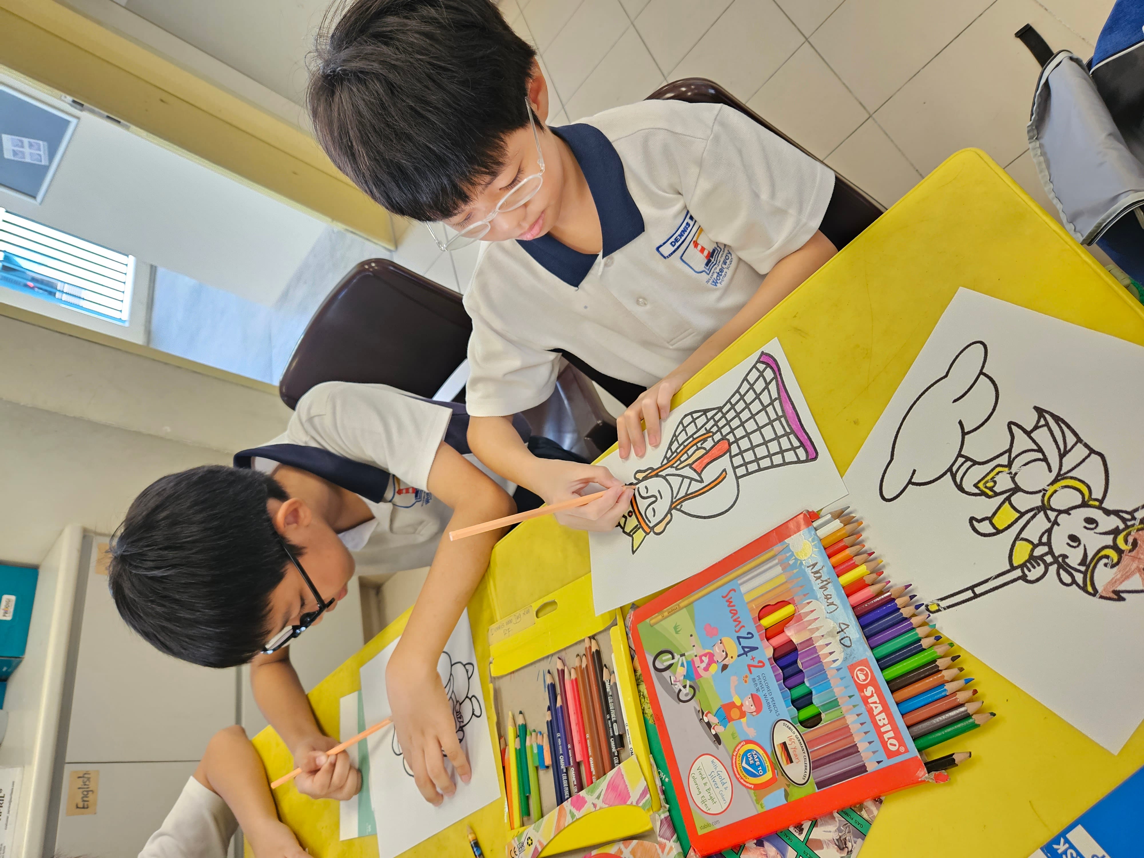 Two young boys wearing glasses color drawings with pencils at a yellow table covered with colored pencils.