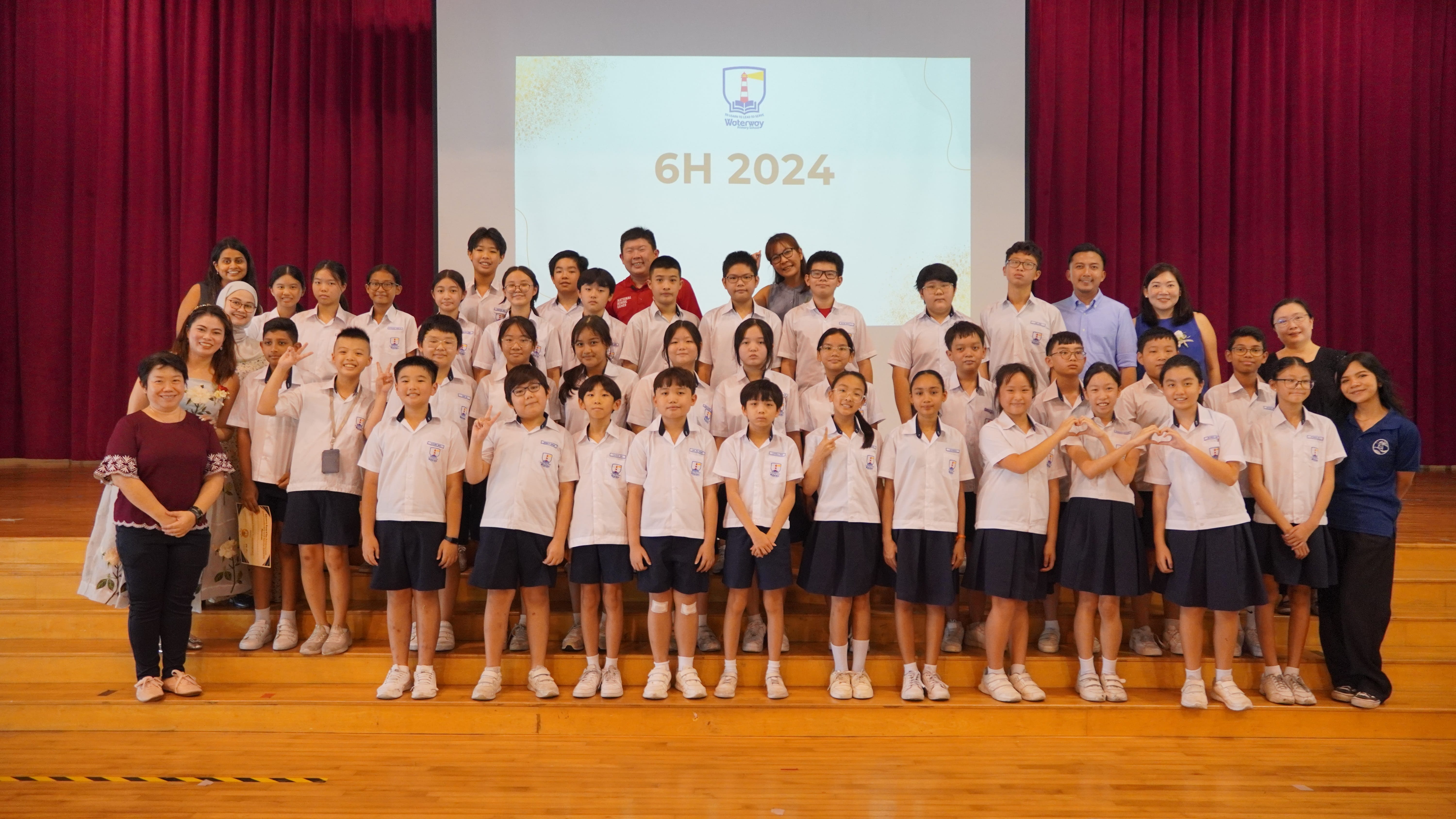 Group photo of students and teachers on a stage; projector screen behind reads 6H 2024.