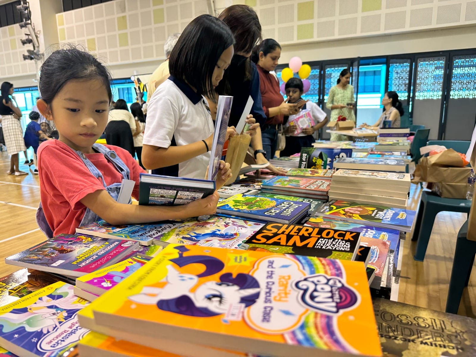 Children and adults browse colorful children's books spread across a table at an indoor book fair.