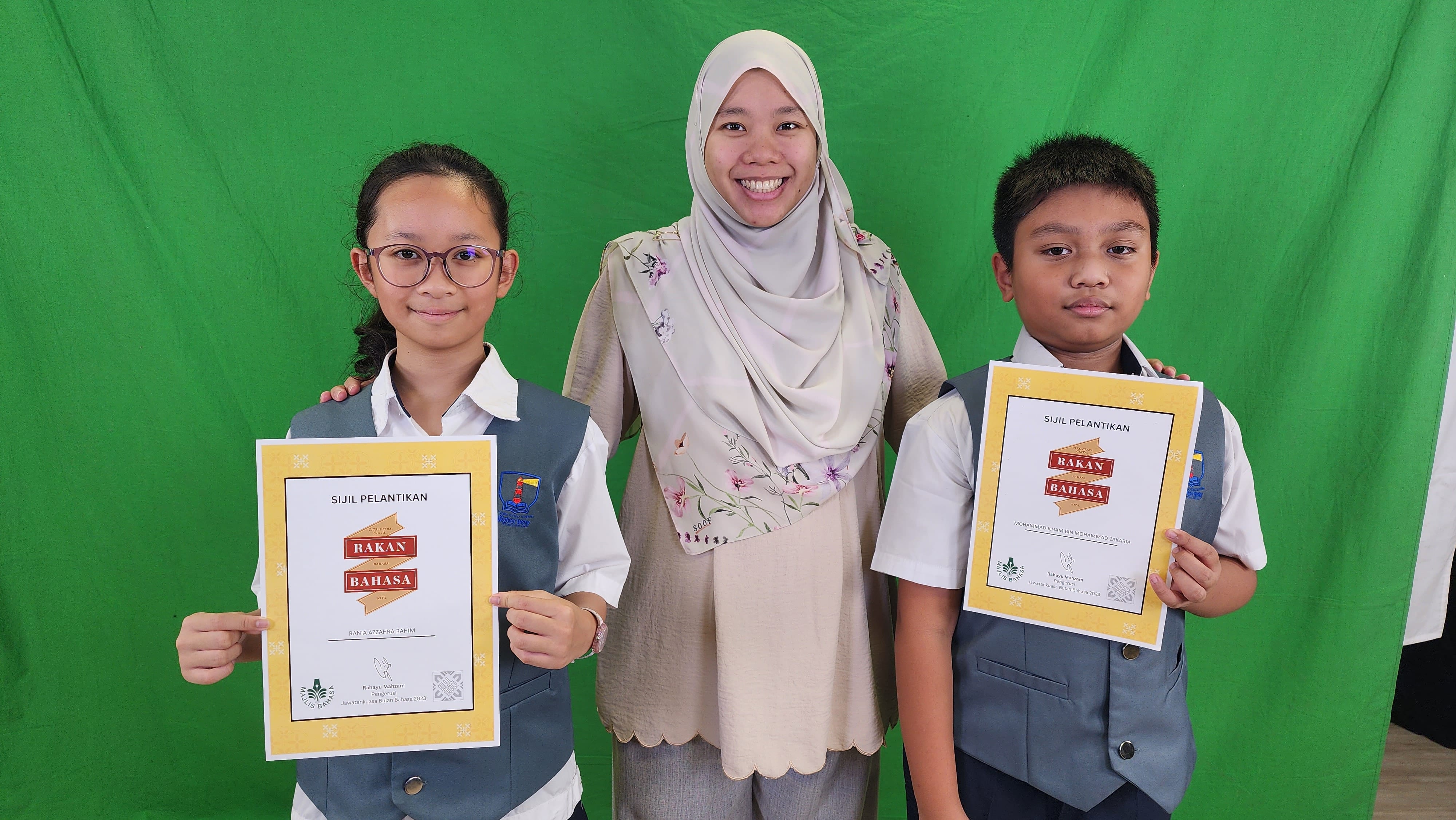 Two students in uniforms hold Sijil Pelantikan Rakan Bahasa certificates, standing with a smiling woman in a hijab.