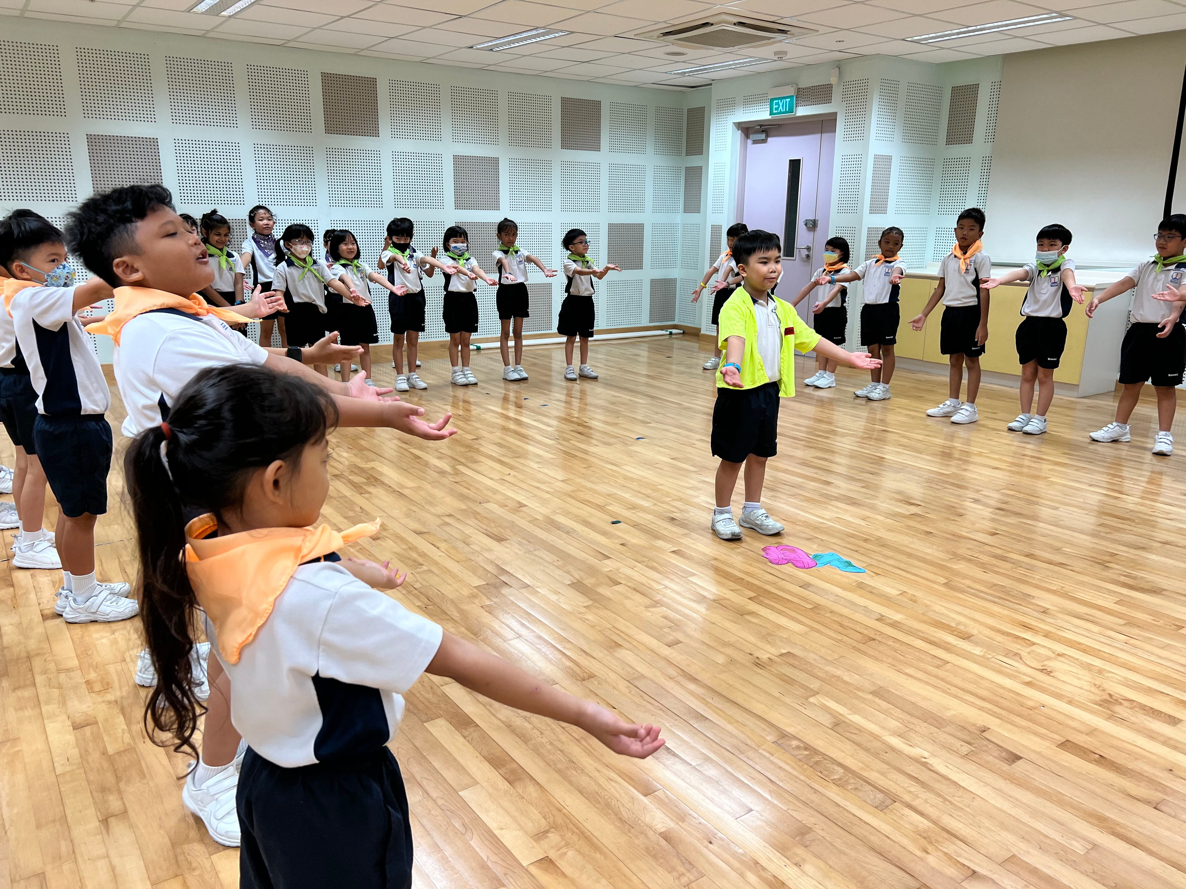 Young school children in uniforms stand in a circle on a wooden floor, stretching arms out during an activity.