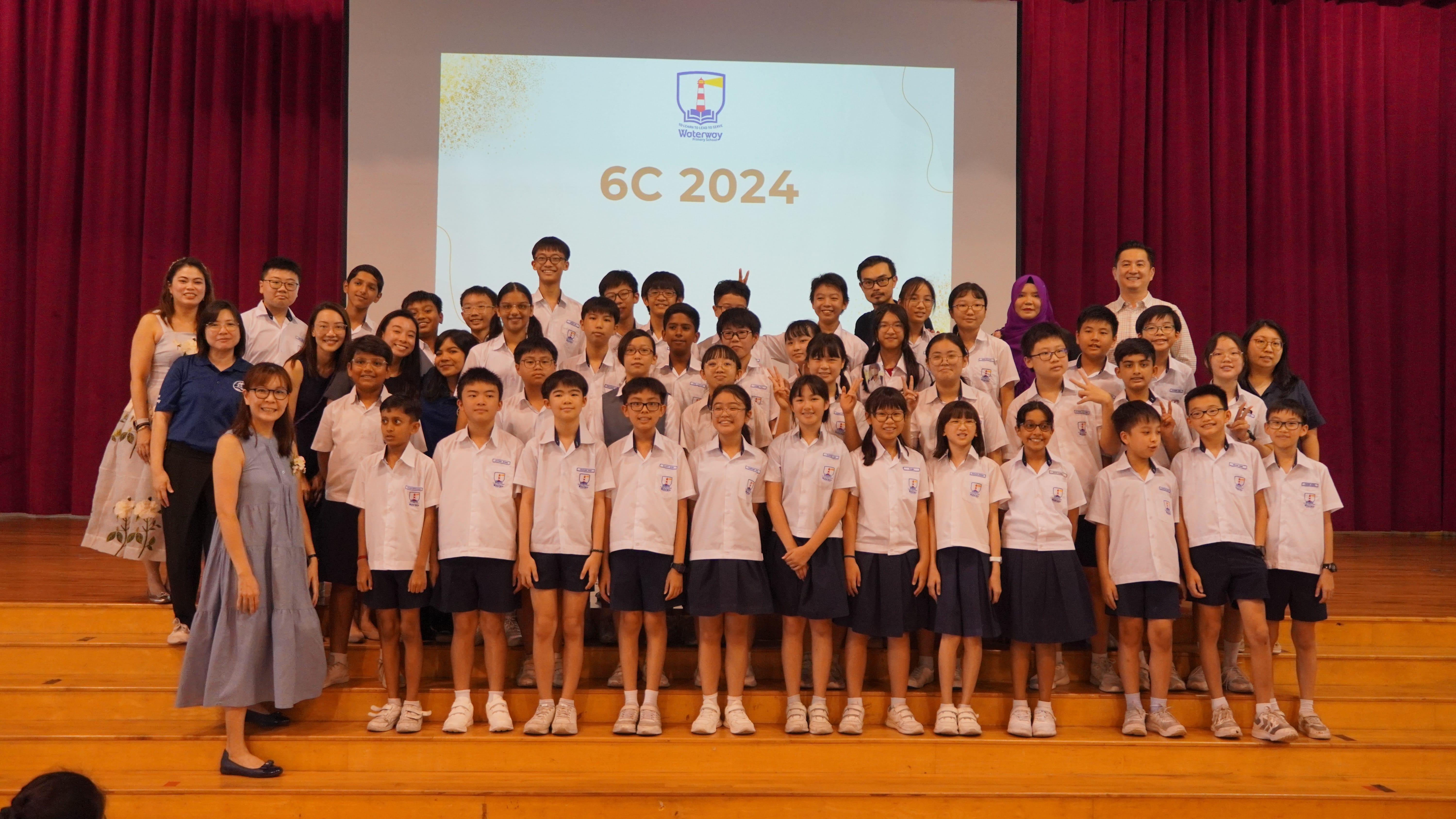 Group of students and teachers in uniforms on a stage, with a screen behind reading 6C 2024.