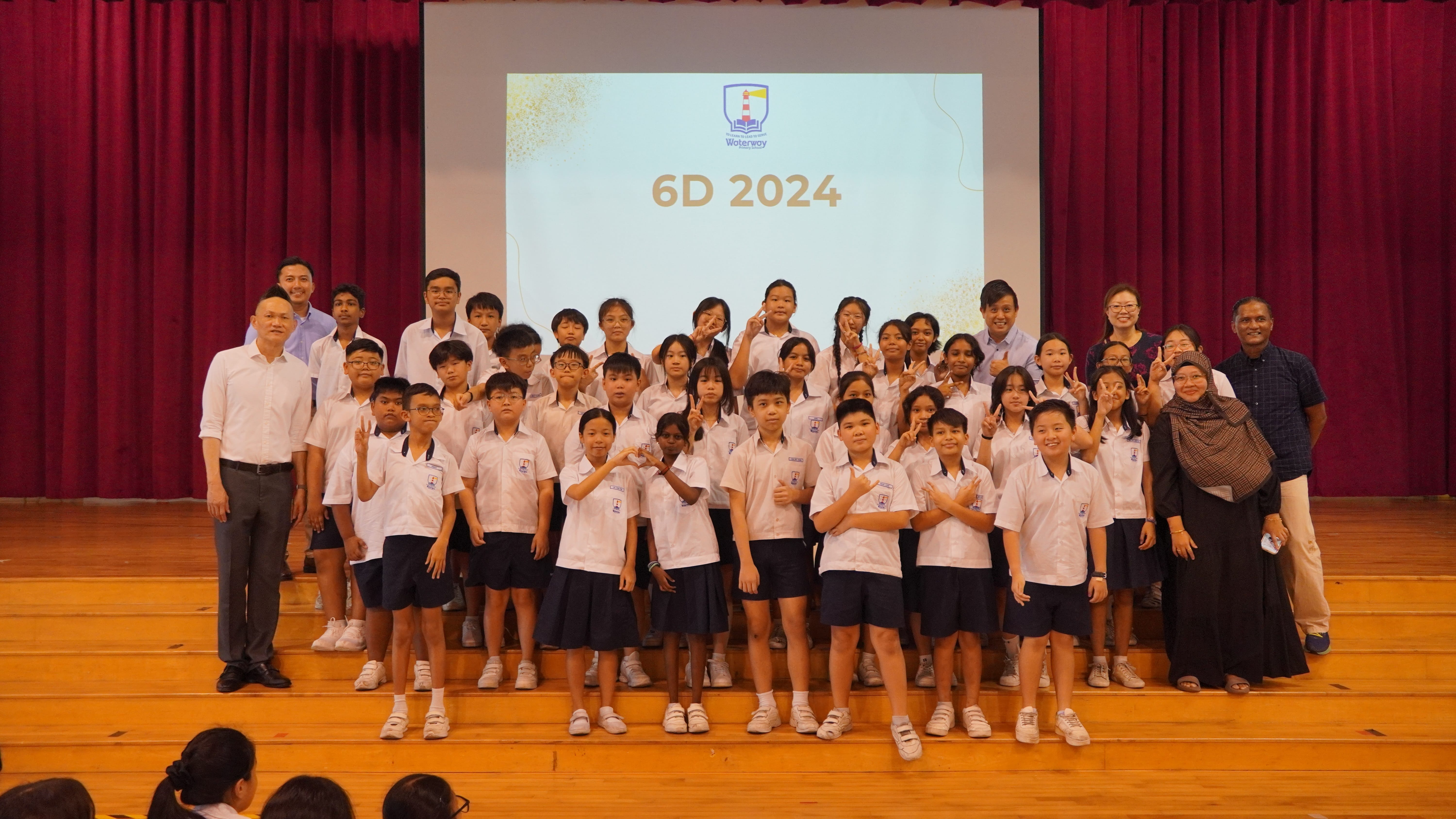 Group of students and teachers in uniforms on a stage, with a screen behind reading 6D 2024.
