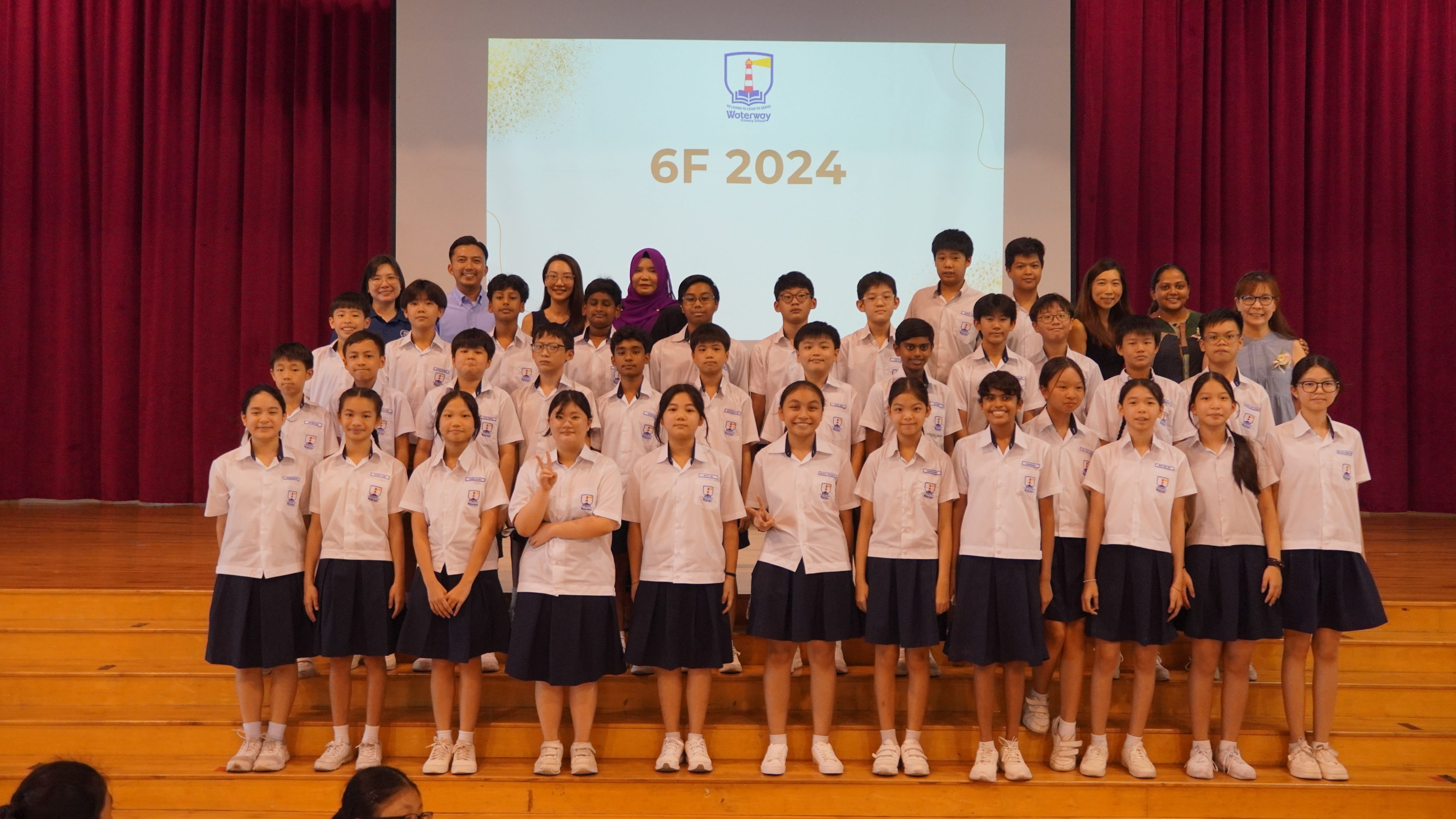 Group of students and teachers in uniforms on stage; screen behind reads 6F 2024, with red curtains.