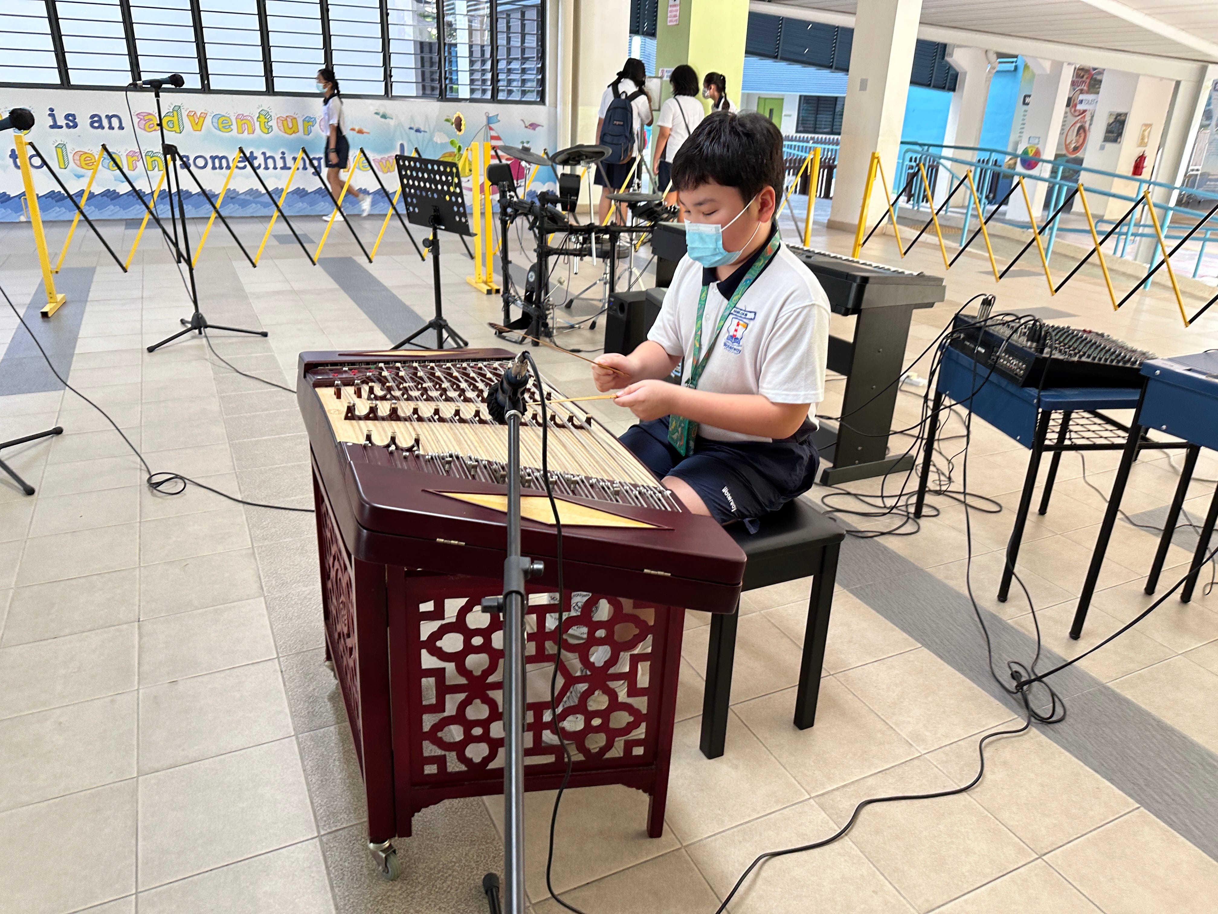 A young boy wearing a face mask plays a yangqin hammered dulcimer in a school hall setting.