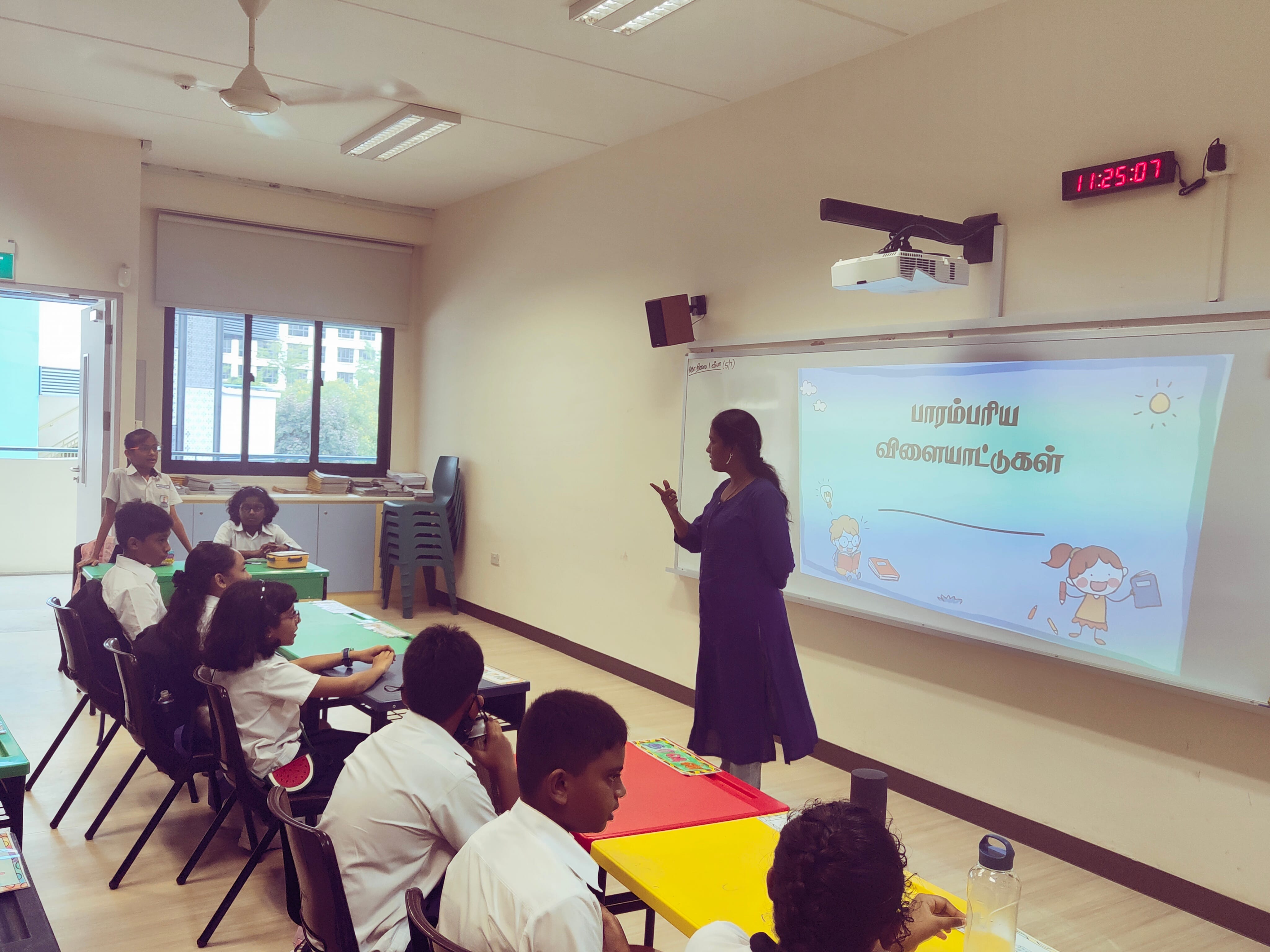 A teacher stands before young students in a classroom, pointing at a projected Tamil-language slide on a whiteboard.