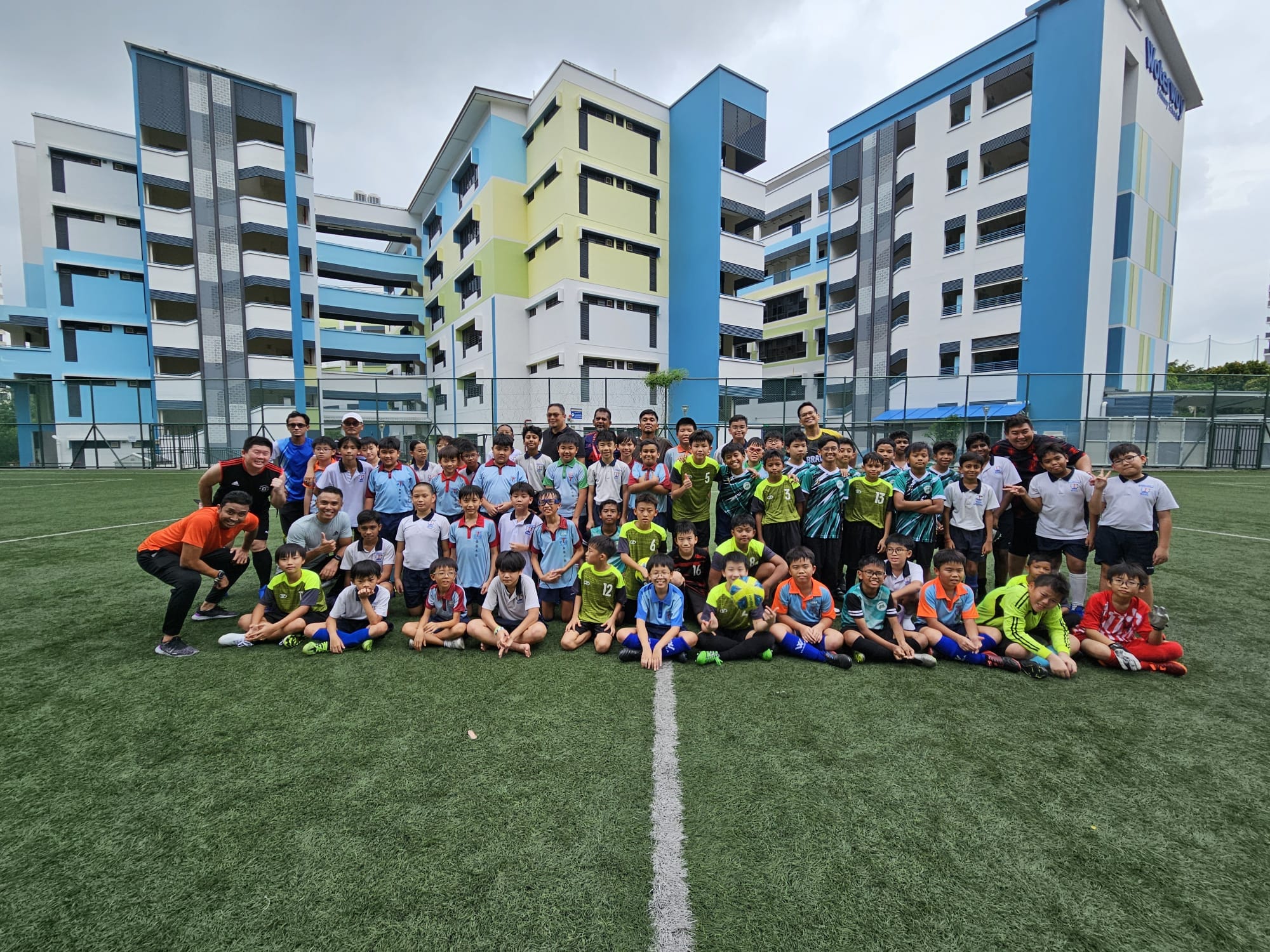 Large group of children and adults in sports jerseys posing on a green football field in front of school buildings.